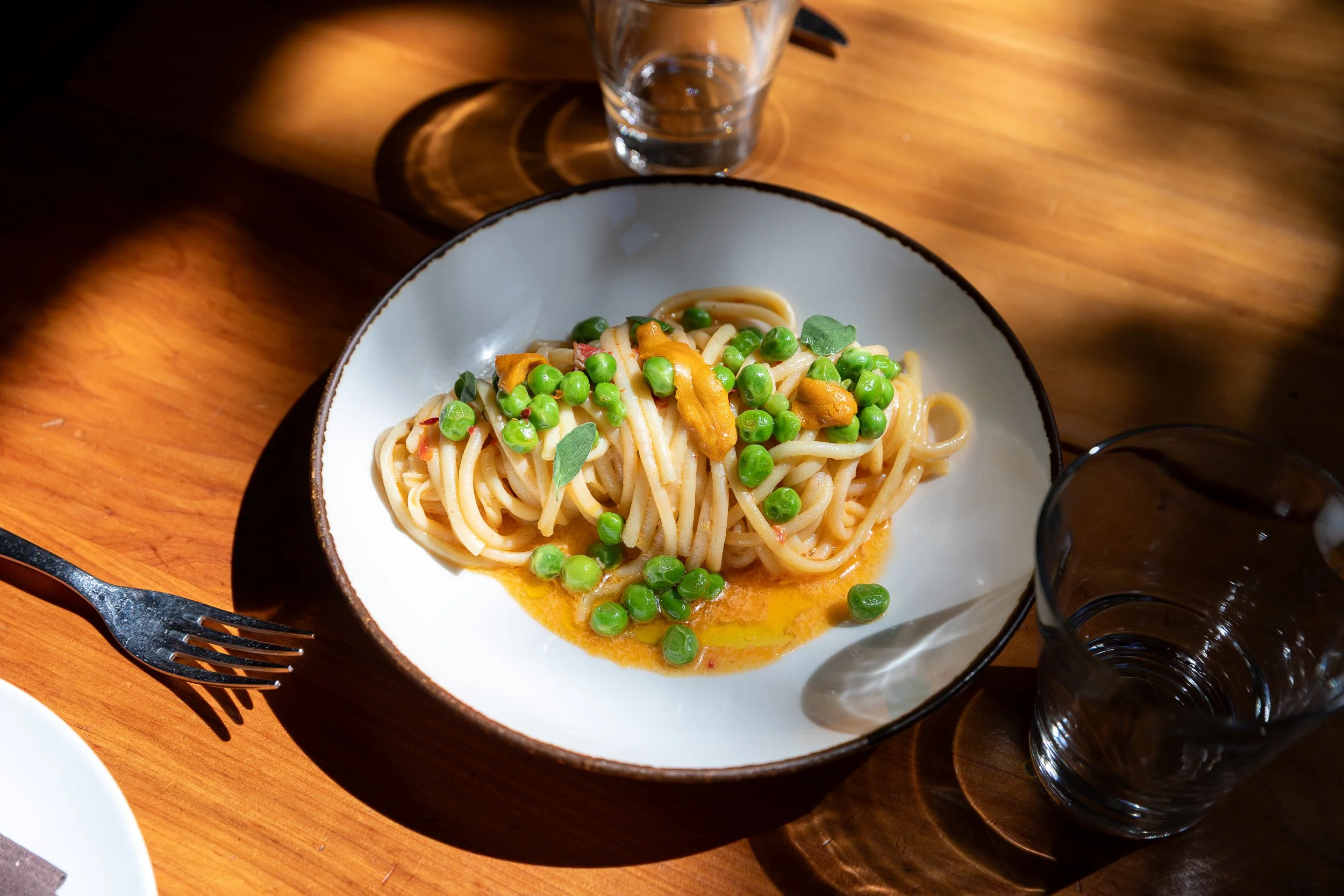 A bowl of pasta garnished with green peas, accompanied by a glass of water on a wooden table.