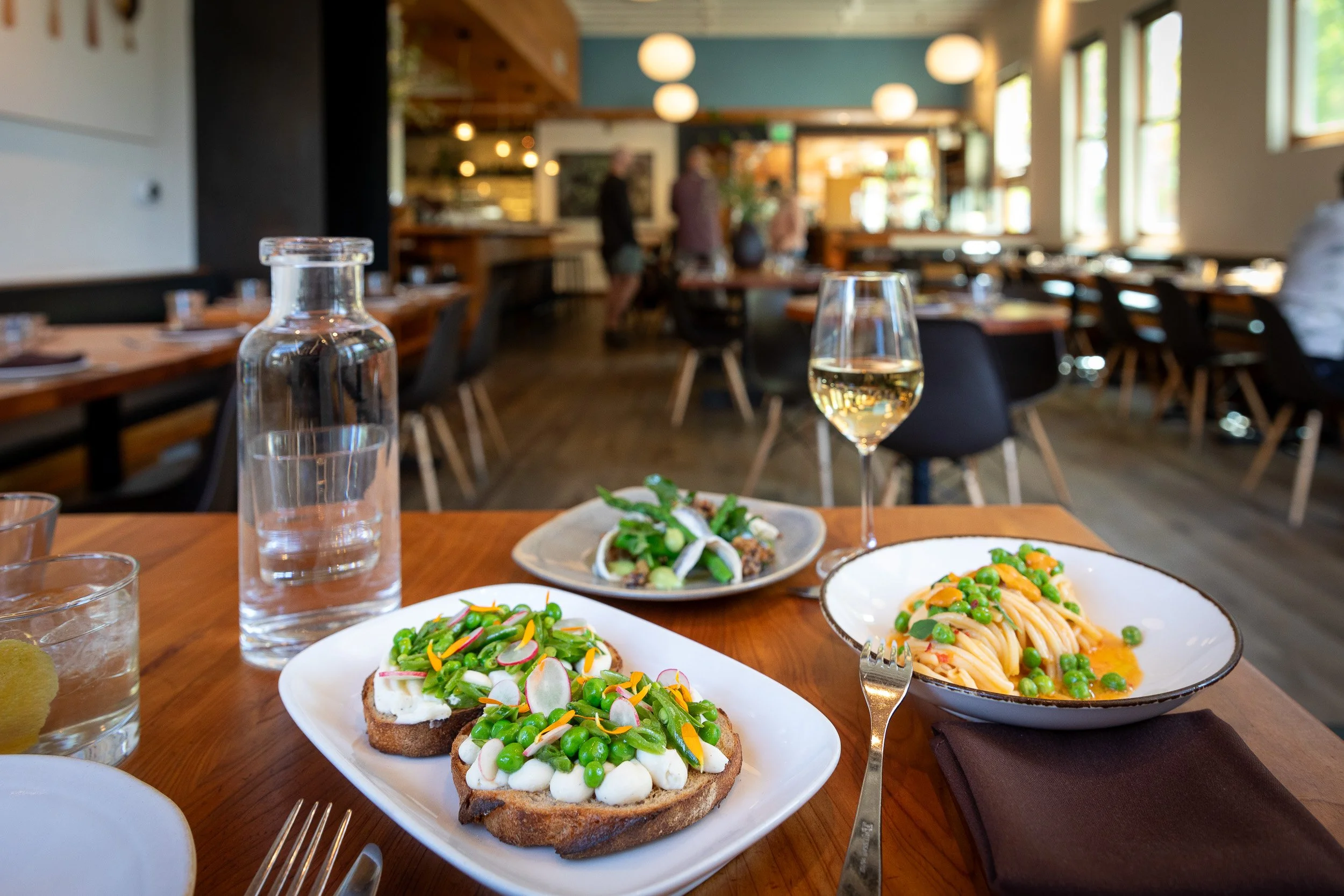 A dining table set with plates of food, including avocado toast and spaghetti, in a restaurant with large windows and blurred background of people and interior decor.