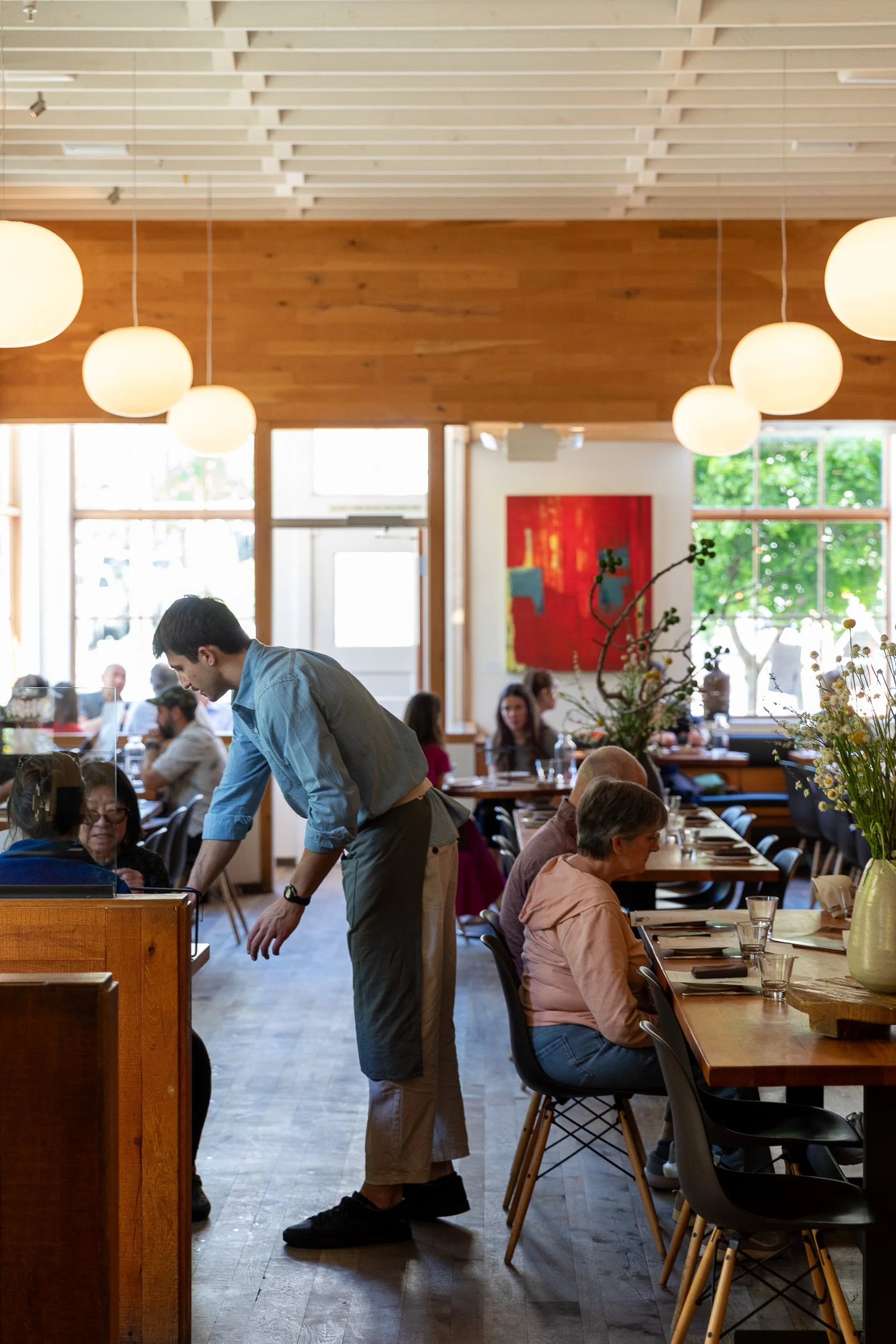 A busy restaurant interior with people dining at tables. A server leans over a table, engaging with a guest. The decor includes wooden elements on the ceiling and a large red abstract painting on the wall. Natural light streams in through large windo