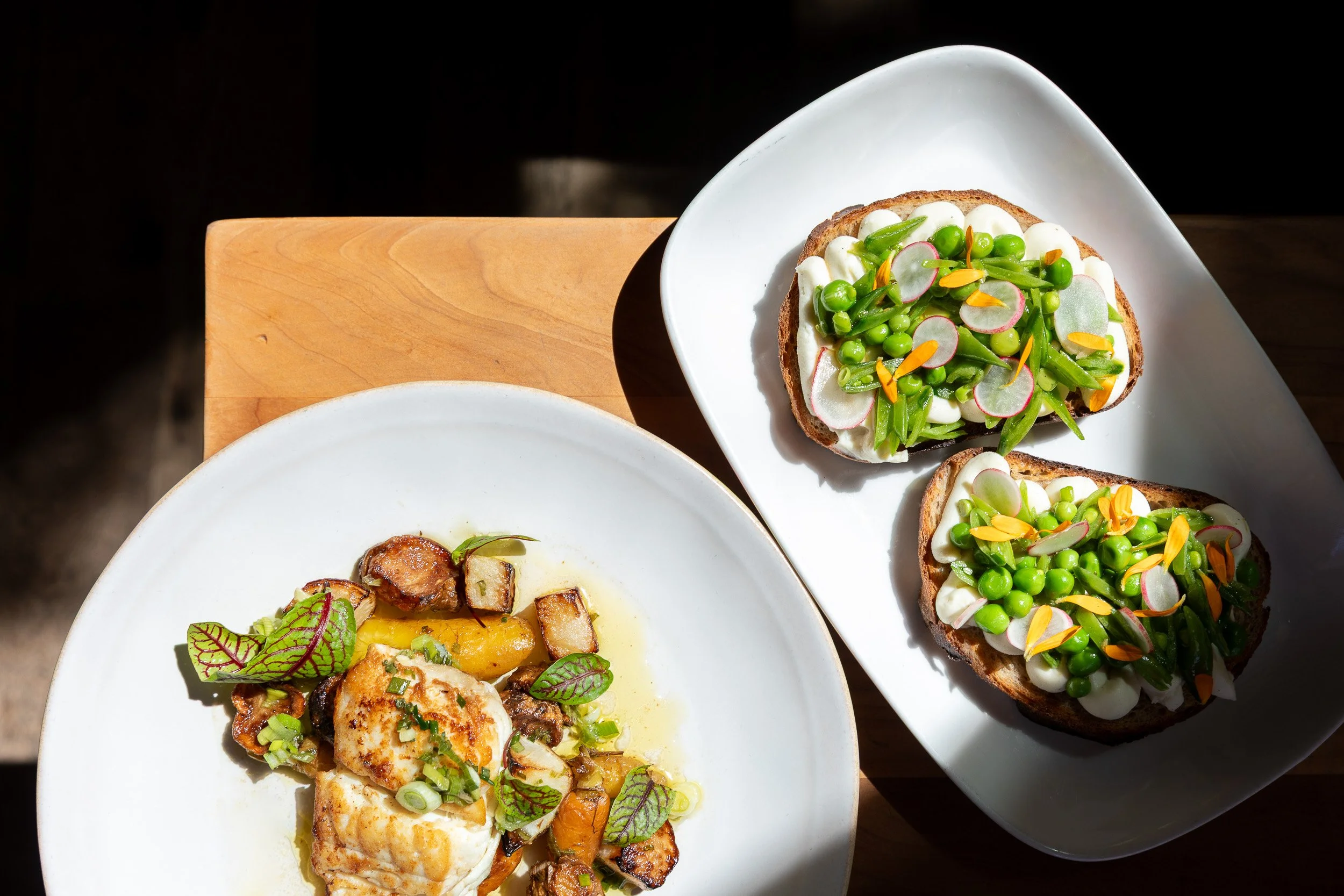 A white plate with grilled chicken topped with chopped green onions and herbs, and roasted potatoes with herbs and green garnish. A white rectangular dish with avocado toast topped with radish slices, green peas, edible flowers, and green onions.