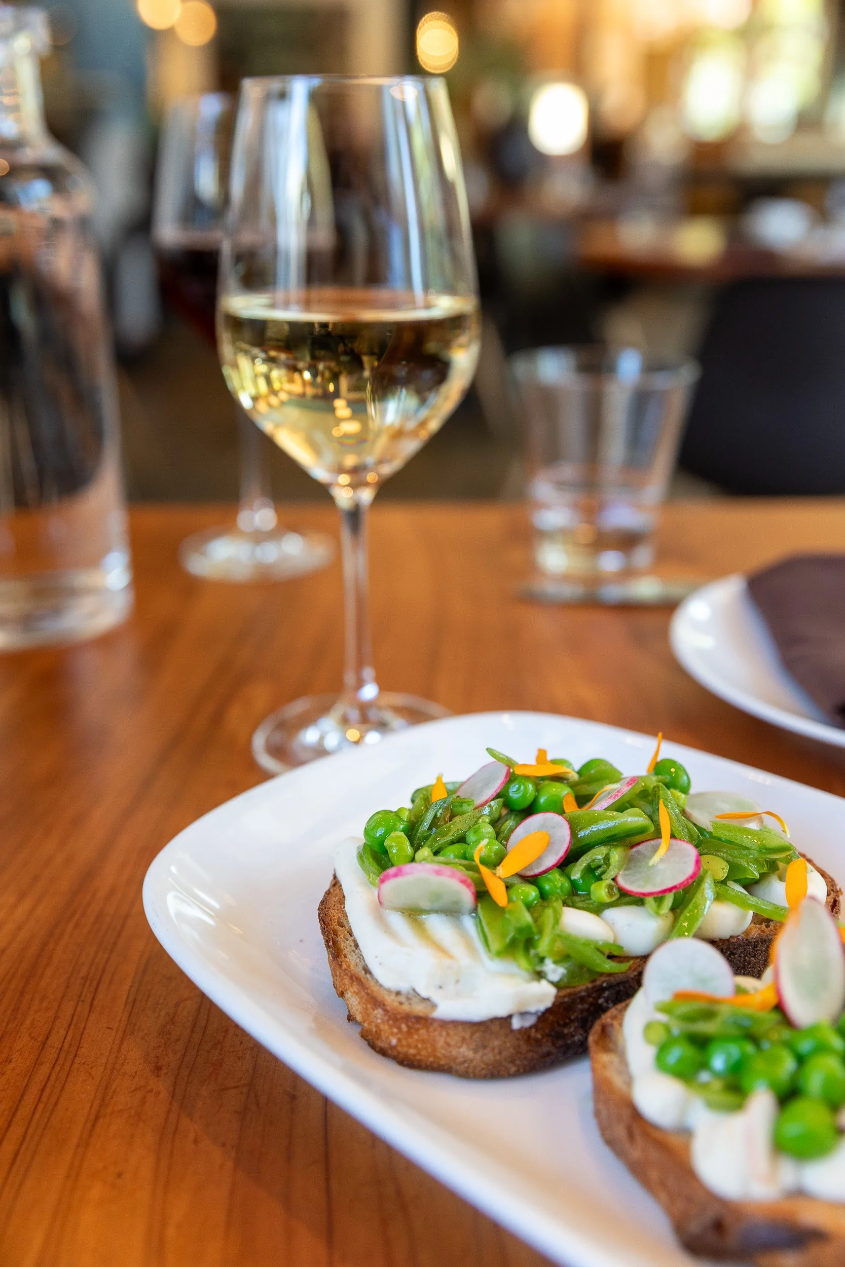 A white plate of open-faced sandwich with creamy spread, topped with green peas, radish slices, and shredded carrots, on a wooden table in a restaurant setting with a glass of white wine and a glass of water in the background.