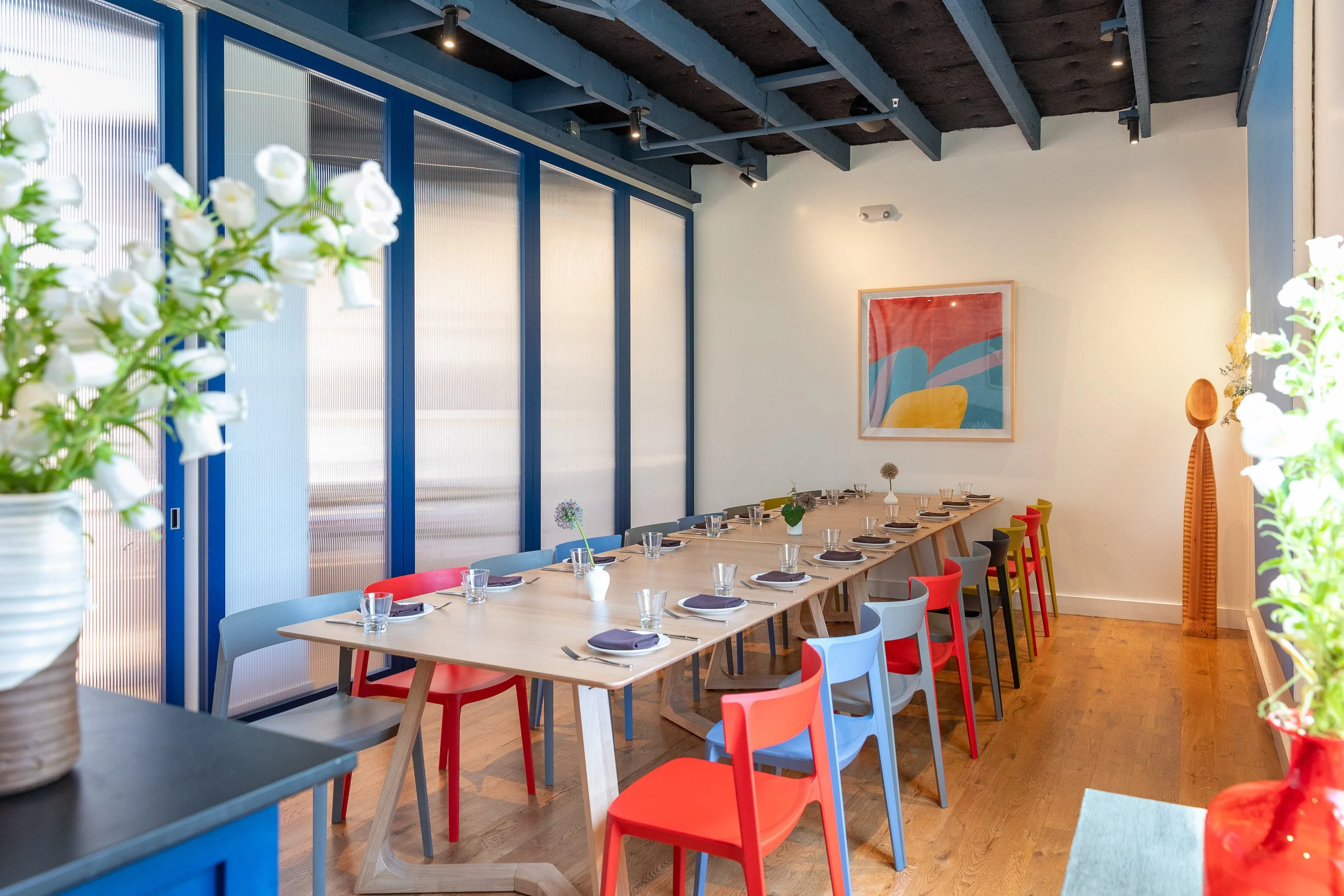 Modern dining or meeting area with a large wooden table, colorful chairs, and decorative vases with greens on the table. There is a blue sideboard, abstract artwork, and an air conditioning unit on the wall. The room has a contemporary design with a mix of vibrant and neutral colors.