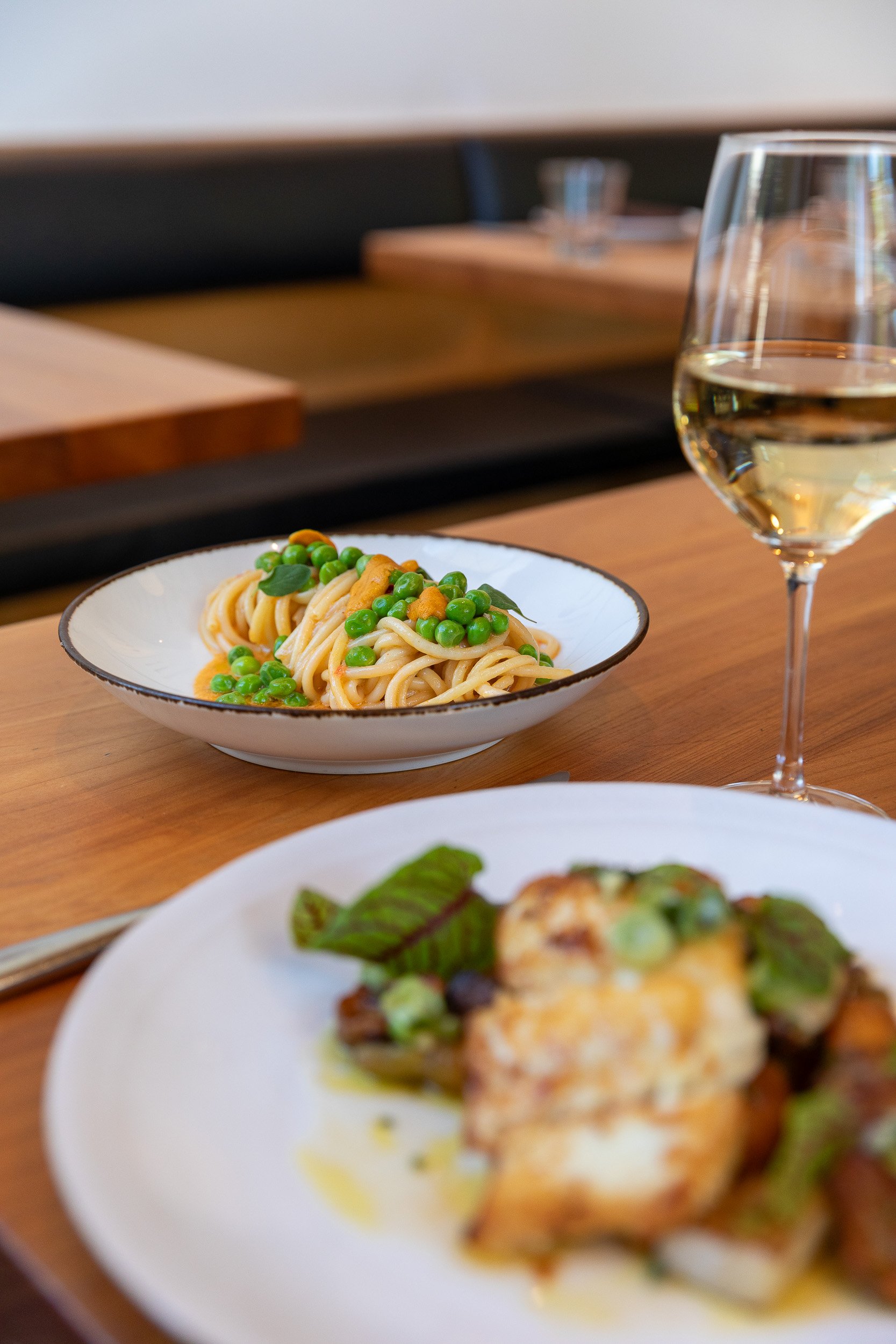 A plate of spaghetti with green peas and possibly some herbs, alongside a glass of white wine, on a wooden table with other blurred dishes in the background.