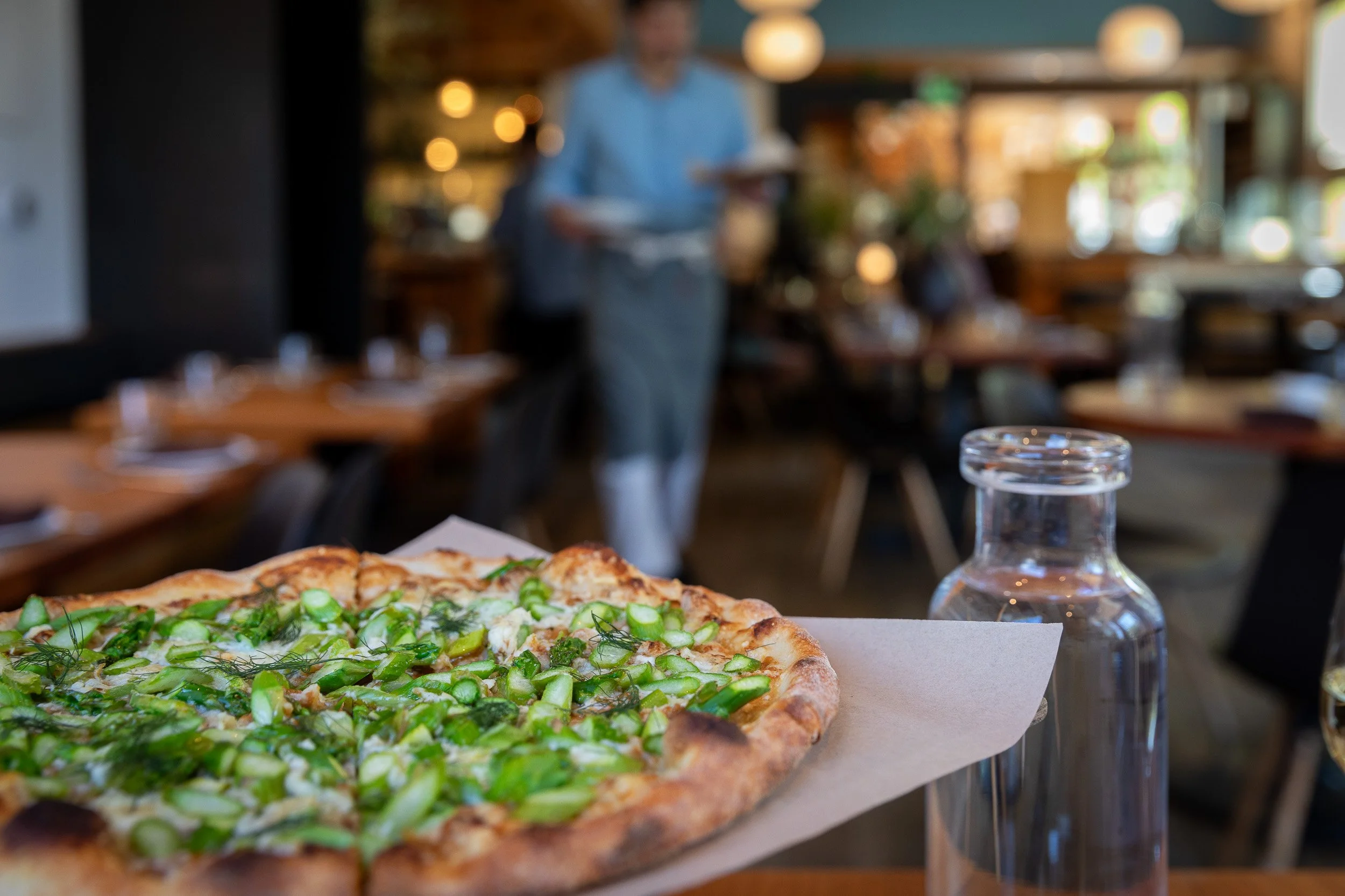 A slice of pizza with green vegetables on top, placed on white paper in the foreground, with a blurred background of a restaurant interior and a person carrying dishes.