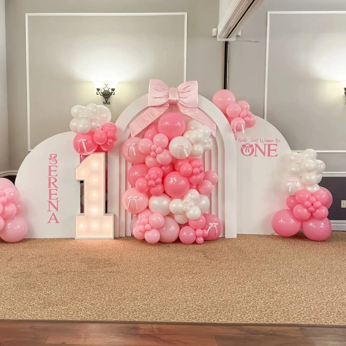 Pink and white balloon decorations for a first birthday celebration with a large illuminated number one, pink ribbon accents, and signs with the names 'Erena' and 'Girls Just Wanna Be One'.