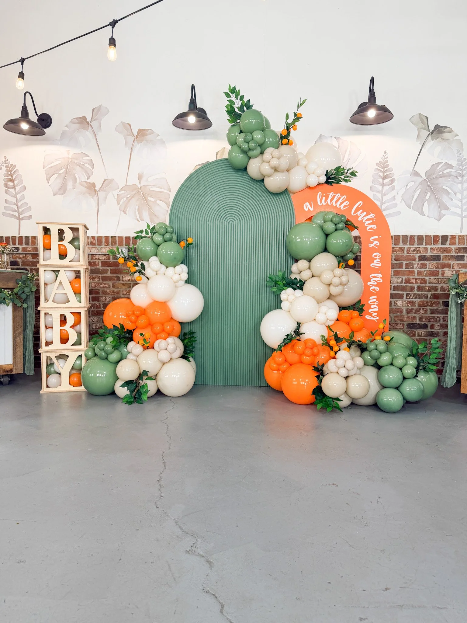 Decorative balloon arrangement with green, white, and orange balloons around a green, semi-circular backdrop. A vertical sign spelling 'BABY' with balloons inside stands to the left. A brick wall with leaf-patterned art and string lights overhead.