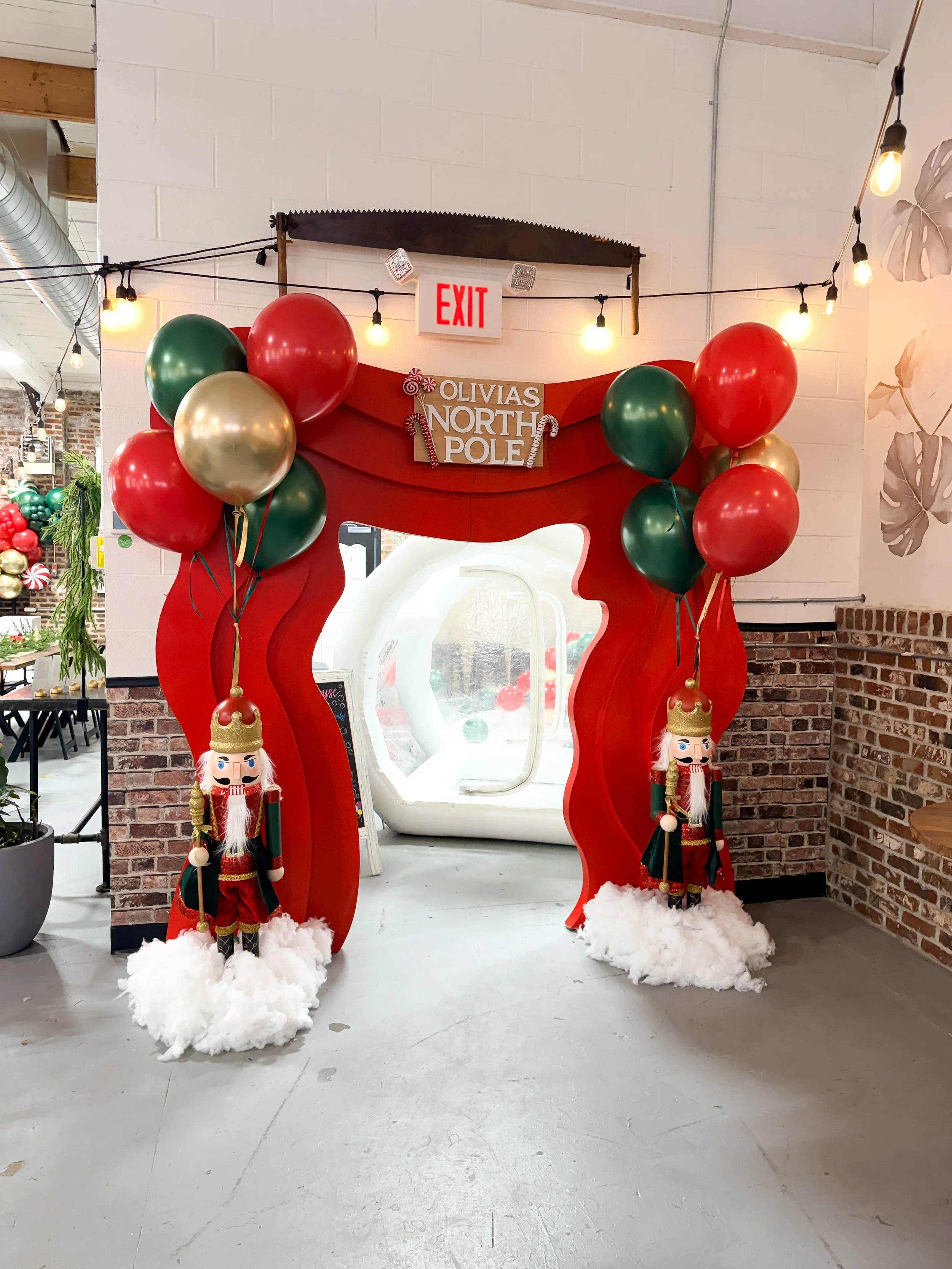 Holiday-decorated doorway with a red frame, green, red, and gold balloons, and nutcracker figures on cotton-like clouds, with a sign that reads 'Olivia's North Pole.'