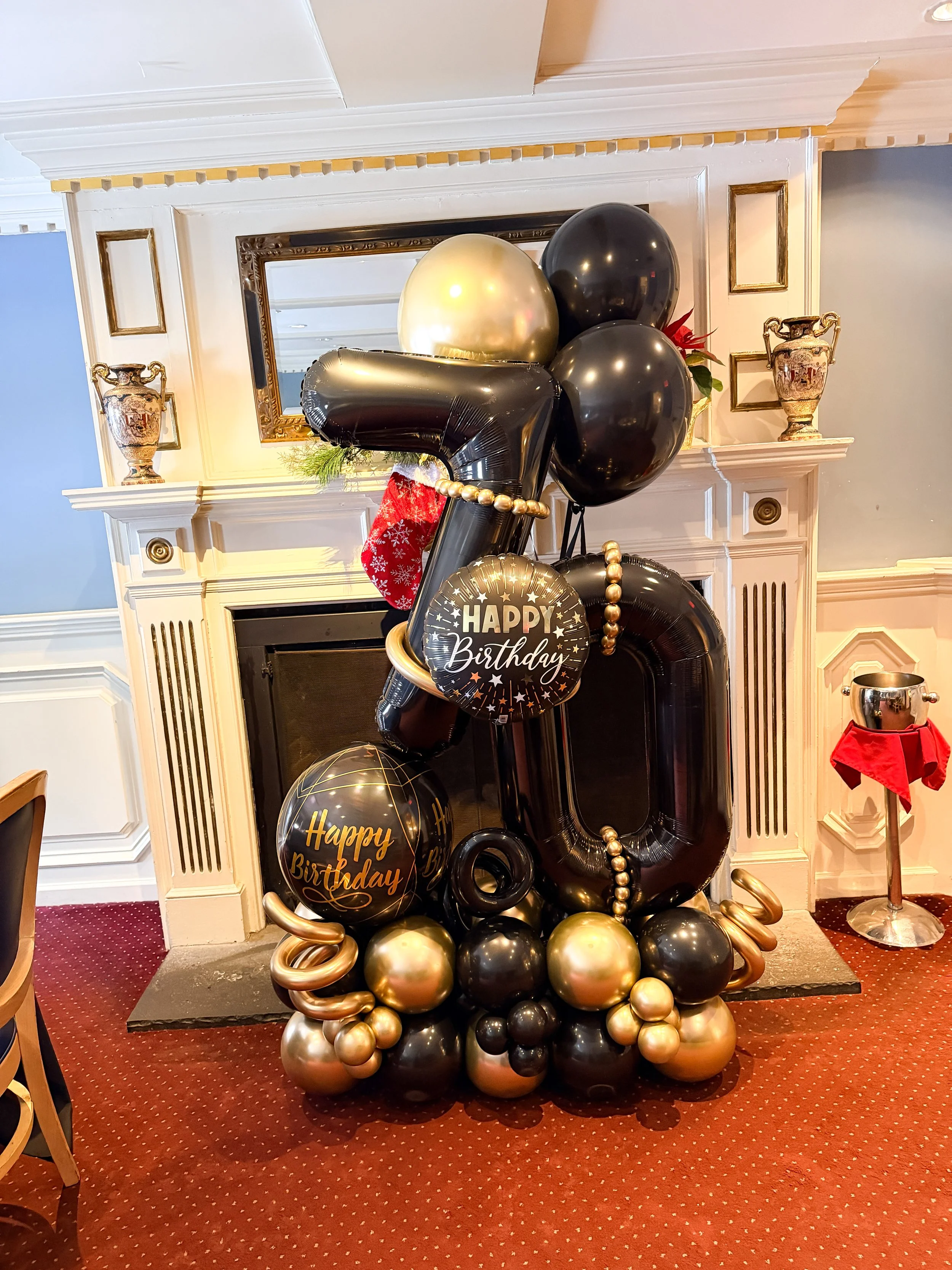 Balloon arrangement with black, gold, and silver balloons, including a large number 70-shaped black balloon, celebrating a 70th birthday at a decorated indoor party venue.