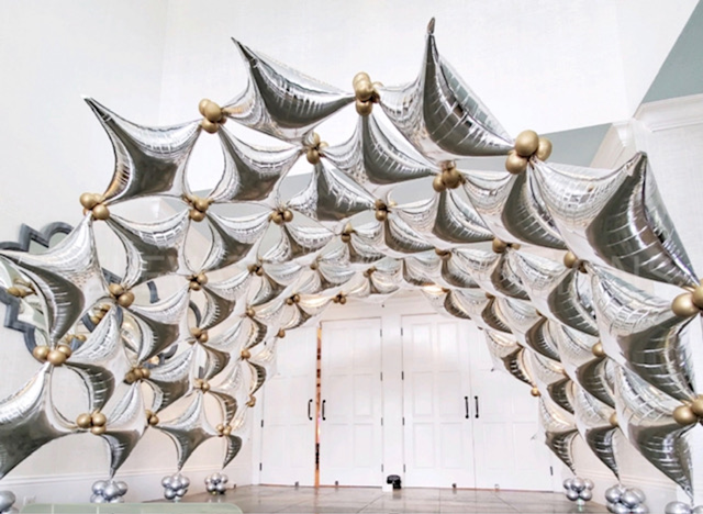Silver and gold star-shaped balloons arranged in an arch inside a room with white walls and doors.
