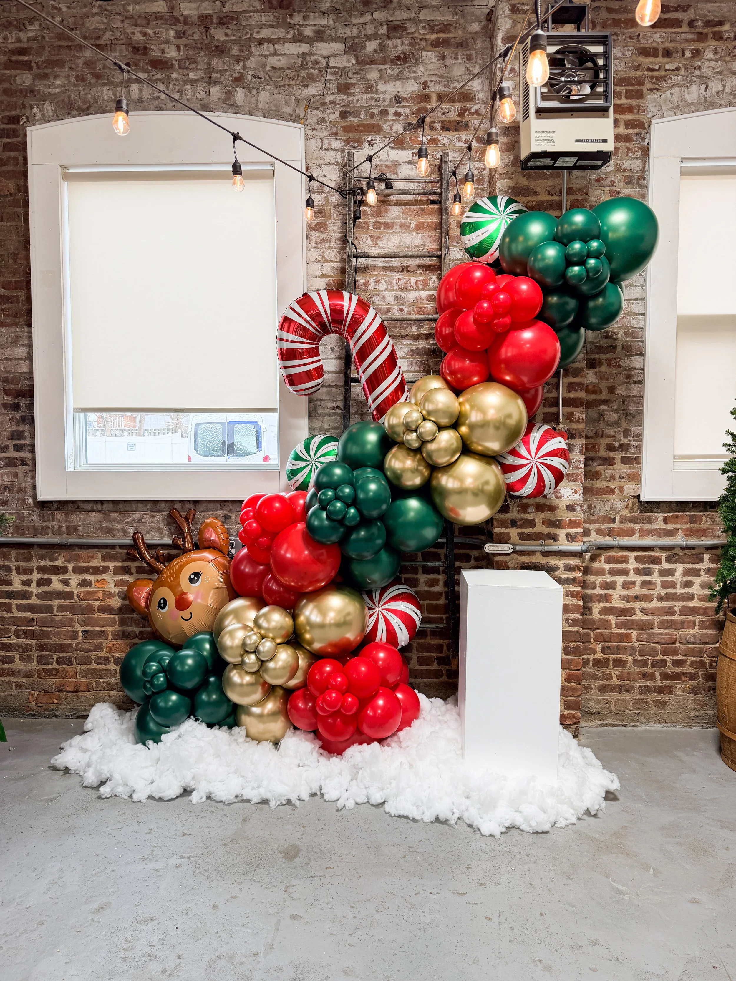 Christmas balloon decoration with red, green, gold, and candy-striped balloons, including a reindeer face balloon, set against a brick wall with windows, string lights, and a white fluffy floor.