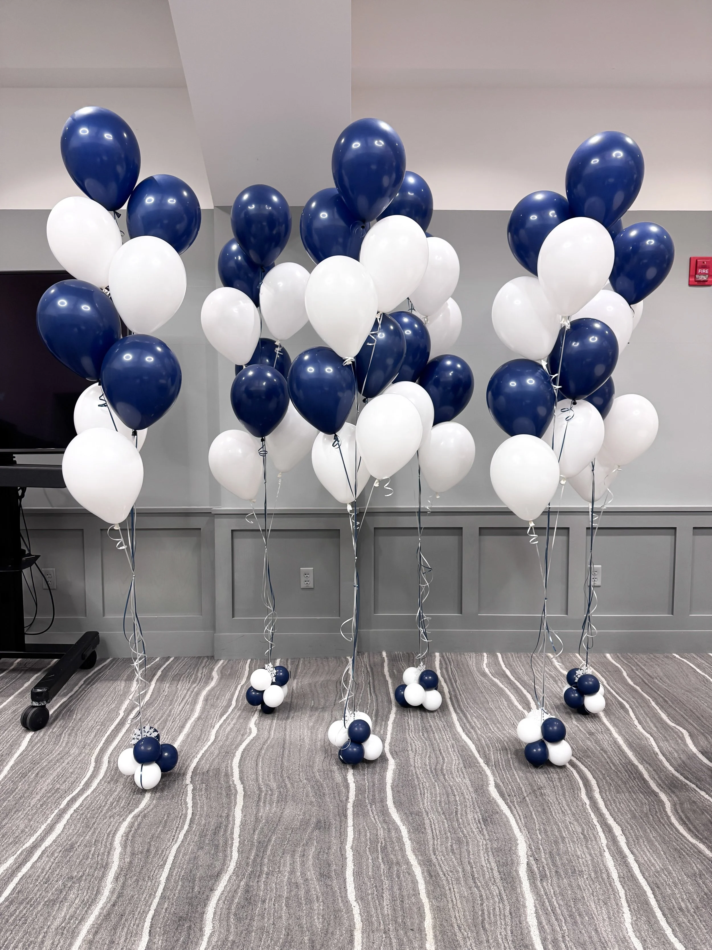 Group of blue and white balloons tied to balloon weights, arranged in clusters on the floor and floating against a plain wall.