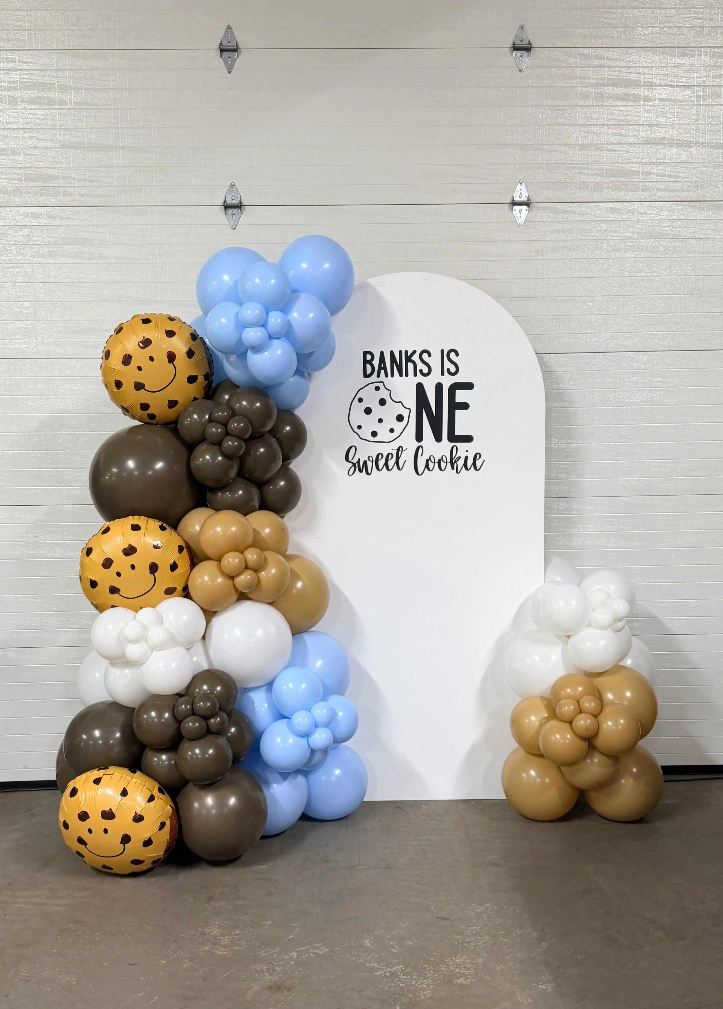 Balloon decoration featuring brown, tan, white, and light blue balloons, with two round balloons with smiley face giraffe pattern, arranged beside a white backdrop with a cookie-themed sign that reads "Banks is one, sweet cookie."