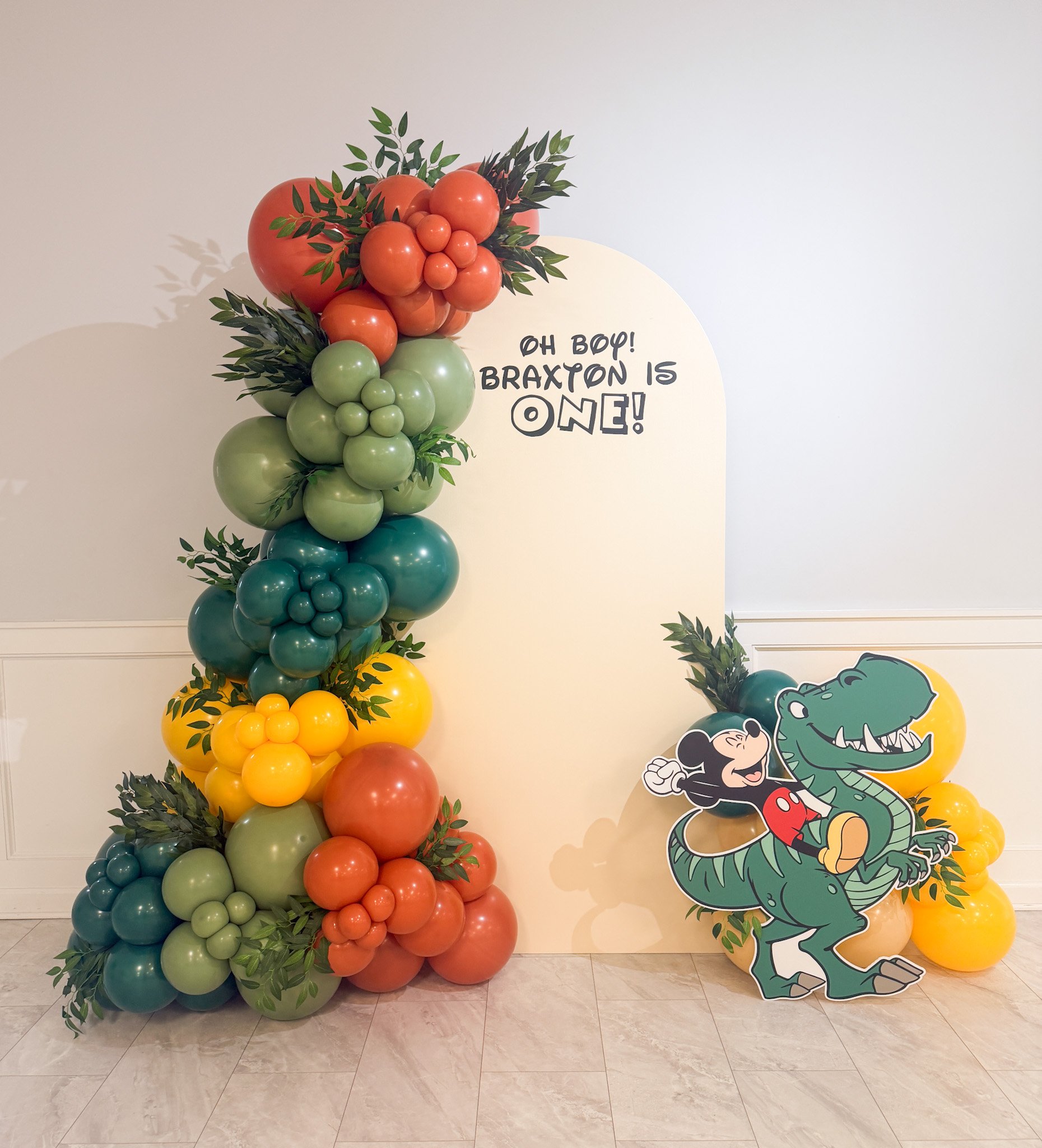 Colorful balloon arrangement with red, green, blue, yellow, and peach balloons, decorated with greenery, and a large cutout of Mickey Mouse riding on a cartoon crocodile. A white backdrop with handwritten text reads, "Oh Boy! Braxton is One!"