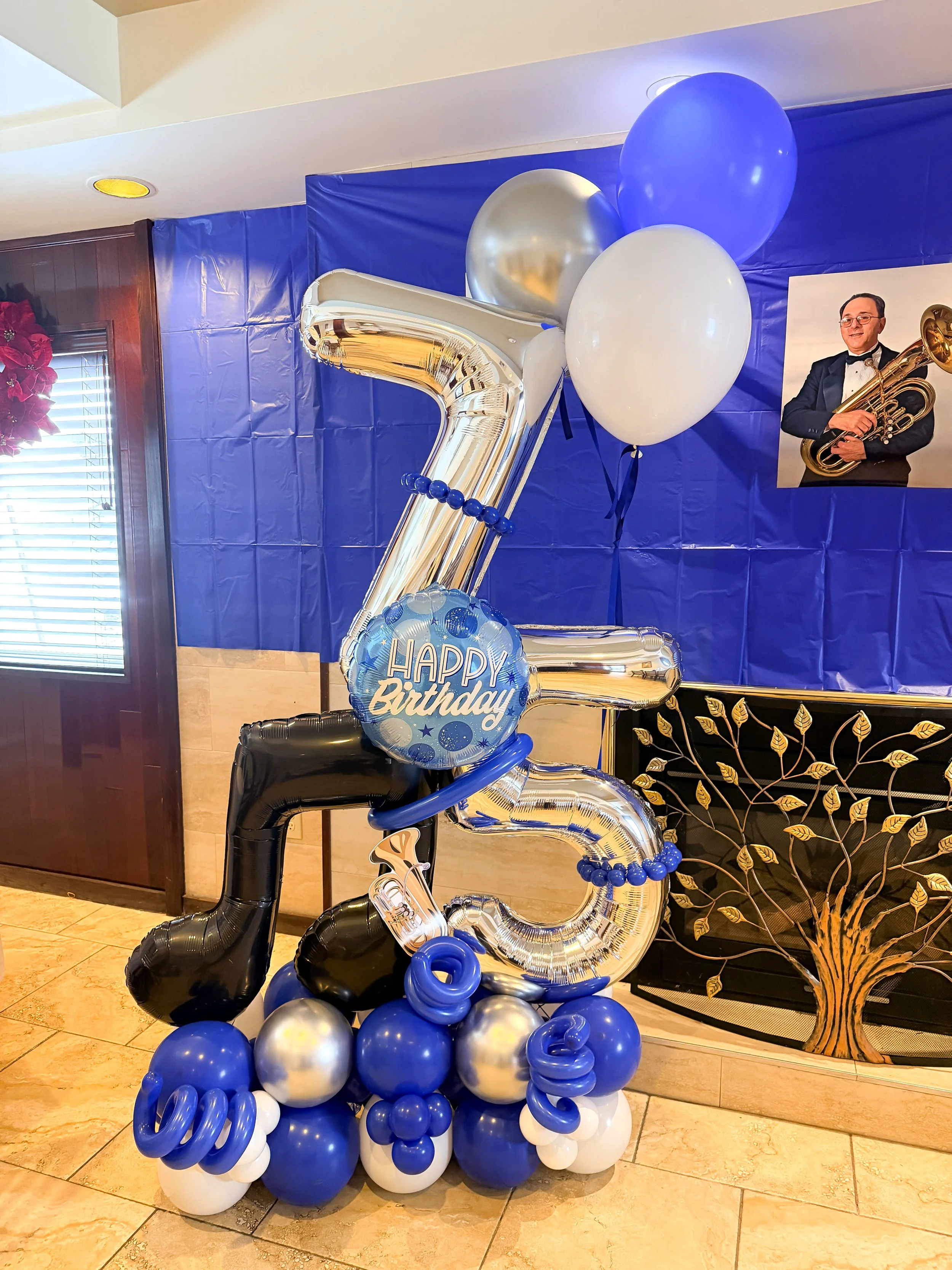 Balloon arrangement celebrating a 75th birthday with large silver and black numbers, blue, white, and silver balloons, and a sign that says "Happy Birthday."