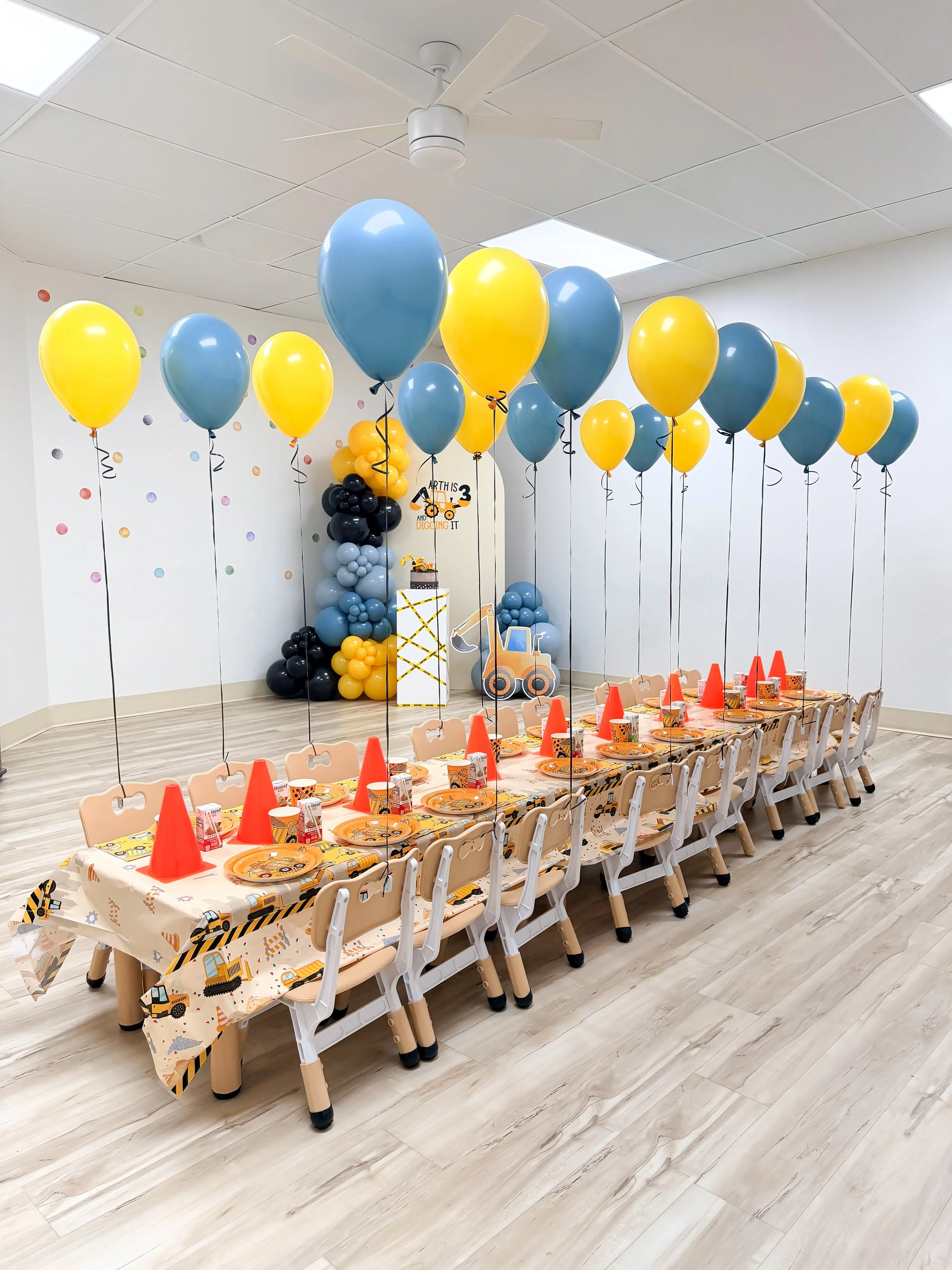 Children's birthday party setup with construction-themed decorations and table settings, featuring yellow and blue balloons, construction vehicle-themed tablecloth, and a cake in the background.