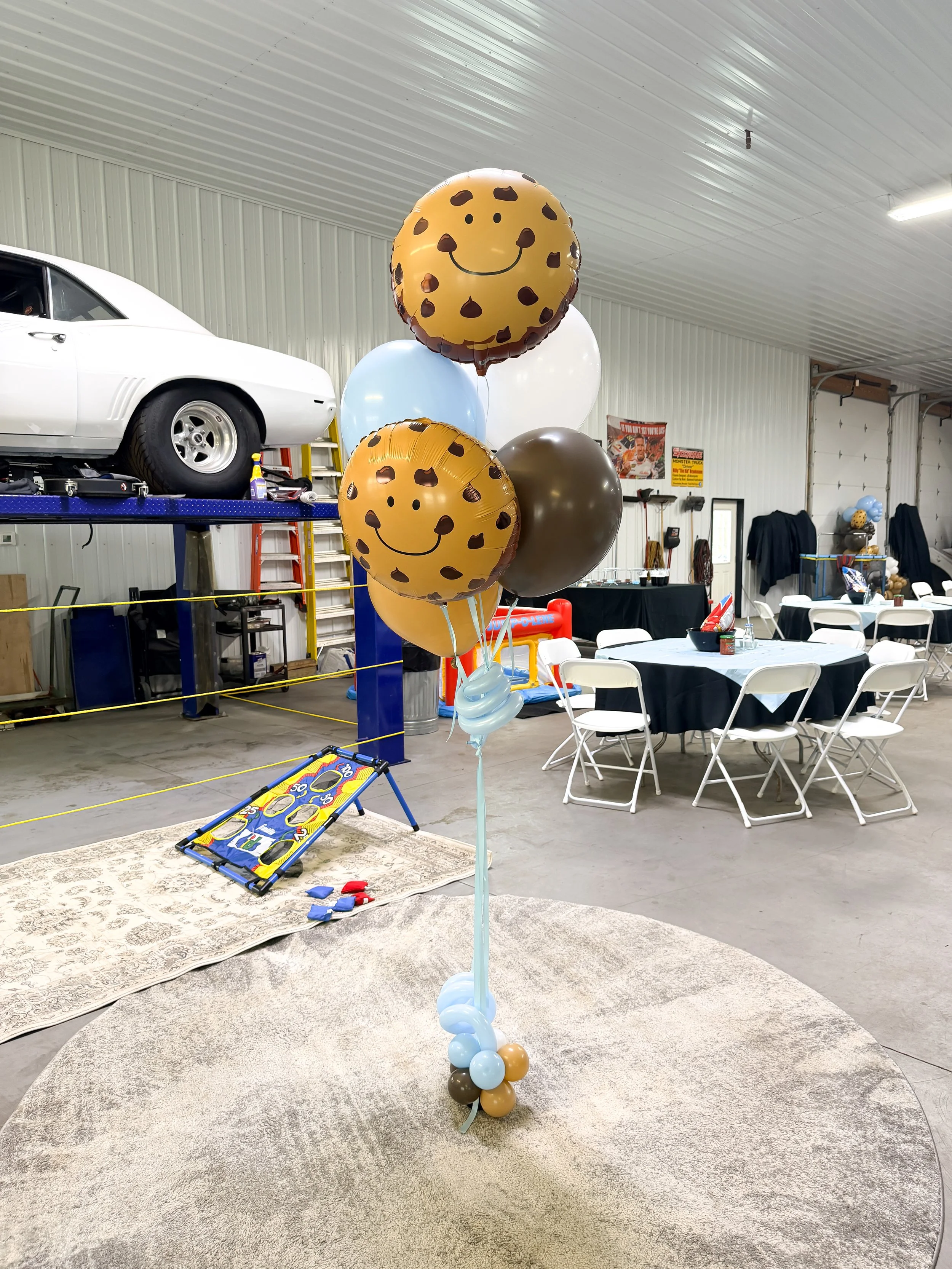 A cluster of birthday balloons, including two smiling cookie-monster patterned balloons, some white, blue, brown, and light blue balloons, and balloons at the base, standing on a round beige carpet in a garage with tables and chairs in the background