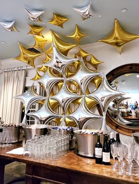 Gold and silver star-shaped balloons decorated on a ceiling and wall, with a table set with wine bottles, glasses, and ice buckets below.