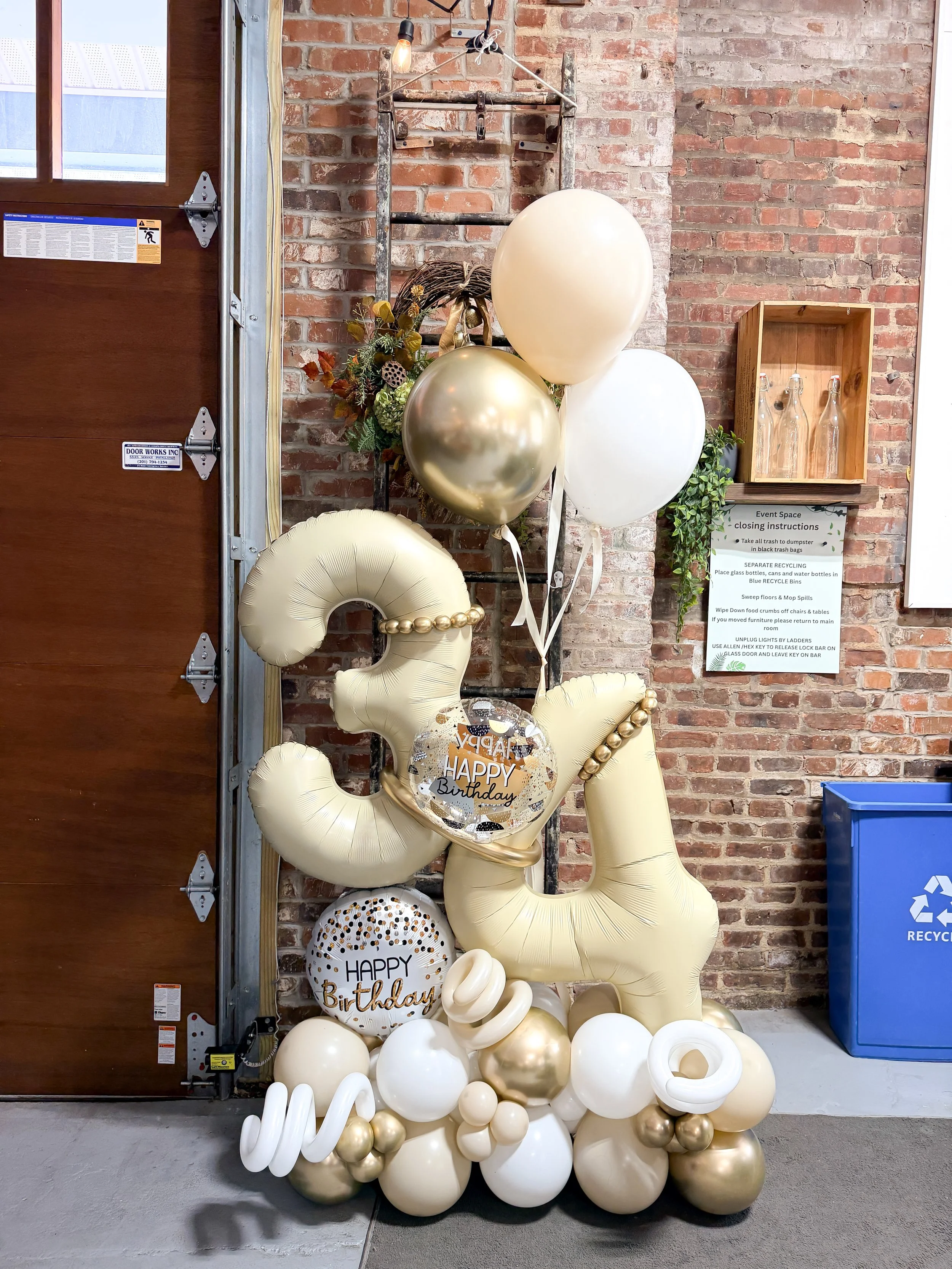 Celebration balloons in gold, white, and cream colors with 'Happy Birthday' messages, arranged with decorative elements against a brick wall.