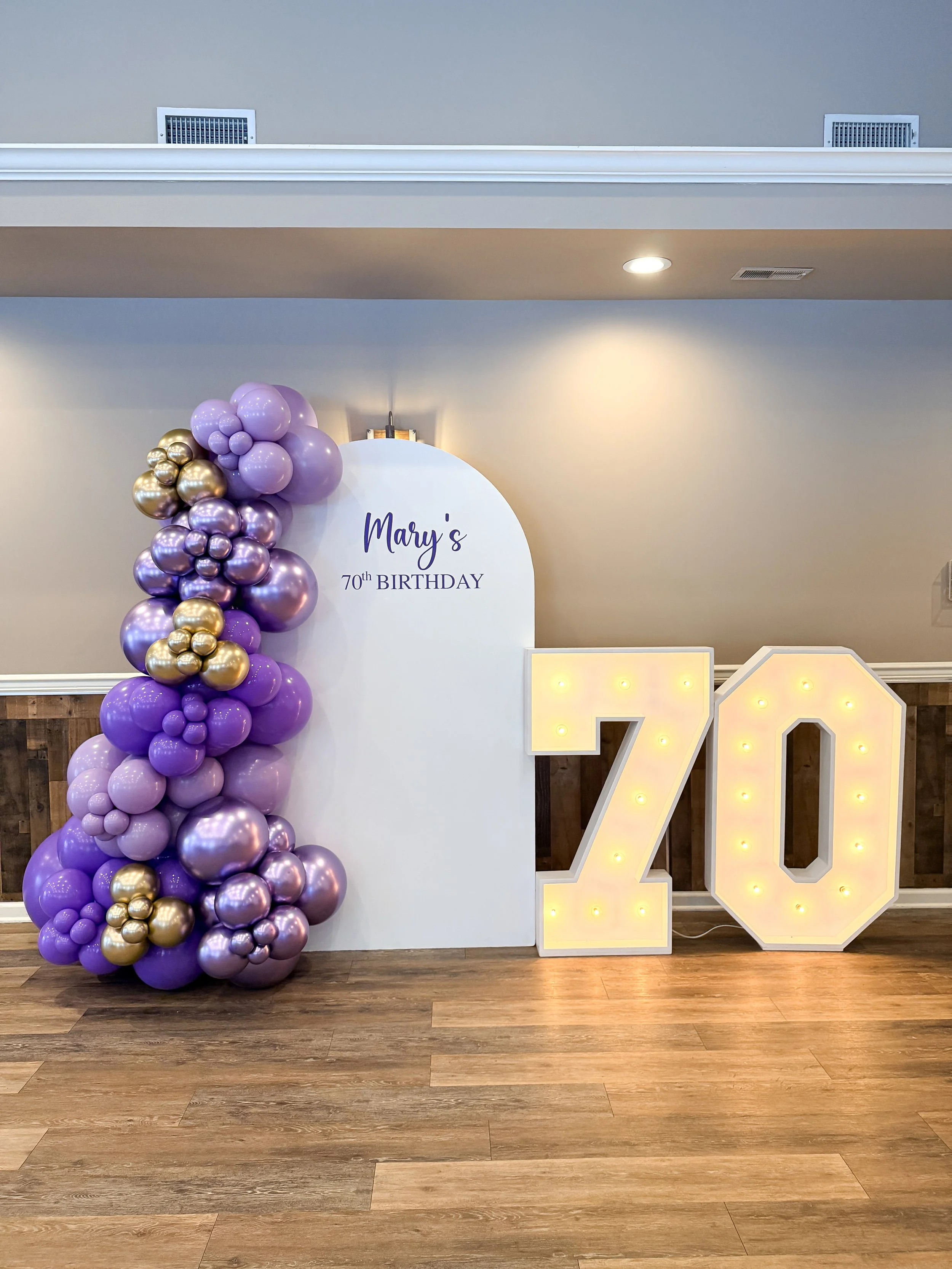 Decorative birthday celebration setup with a white backdrop that says "Mary's 70th Birthday" in purple script. To the left, there is a large arrangement of purple, lavender, and gold balloons. To the right, there are large illuminated numbers "70" in