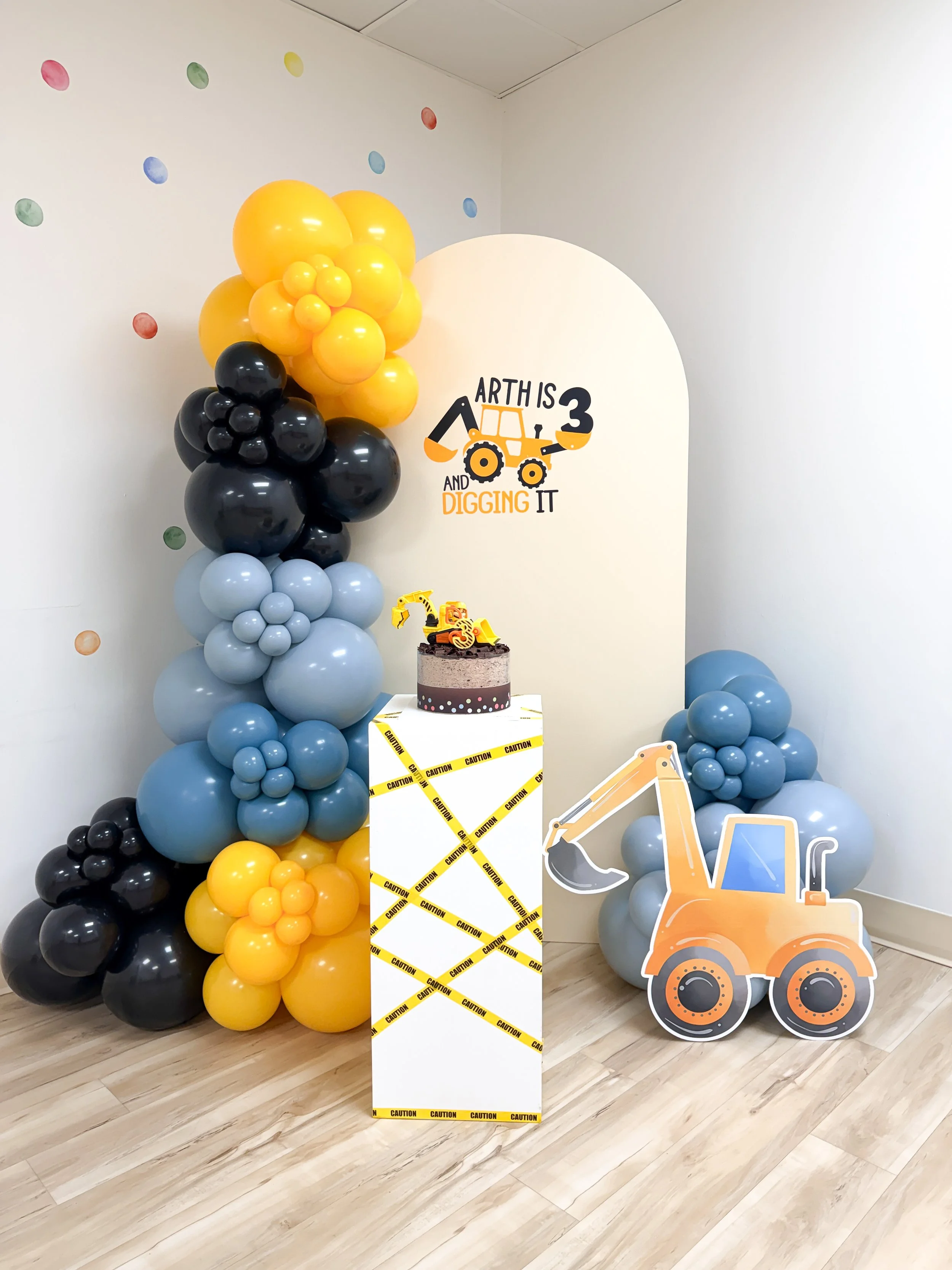 Construction-themed birthday party decorations with a balloon arrangement of yellow, black, and gray balloons, a birthday cake with a construction vehicle topper, a cardboard cutout of an excavator, and a wall sign that reads "Arth is 3 and digging i