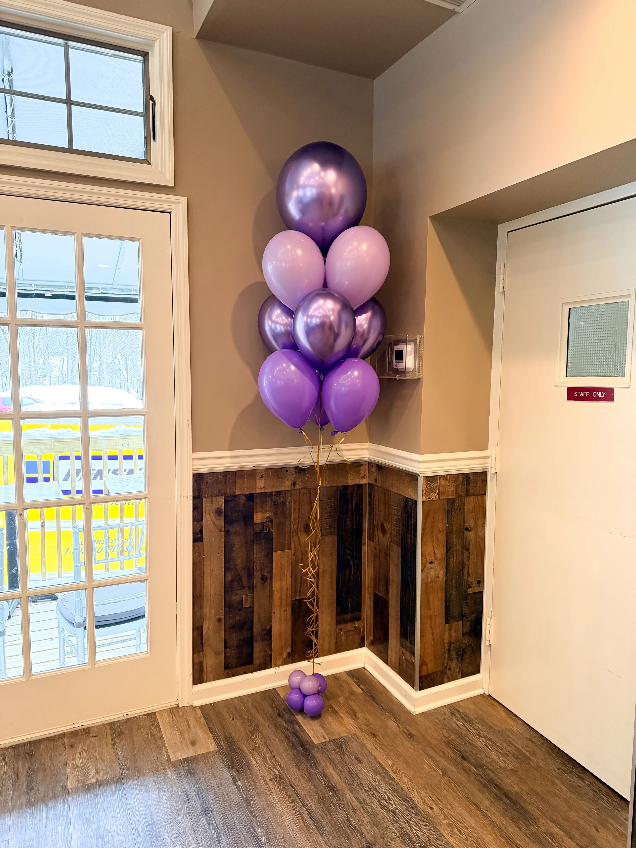 A bunch of purple and lavender balloons tied together with curling ribbons, placed indoors near a white door with a small window and a waiting area visible outside through a glass door.