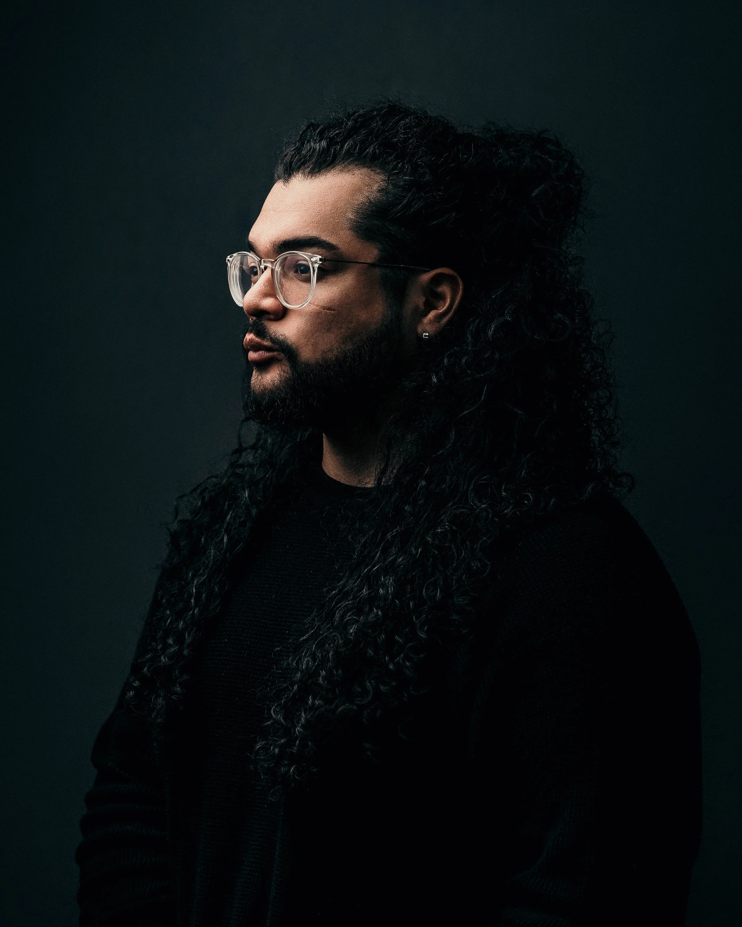 Side profile of a man with long curly hair, glasses, and a beard, wearing a black shirt against a dark background.
