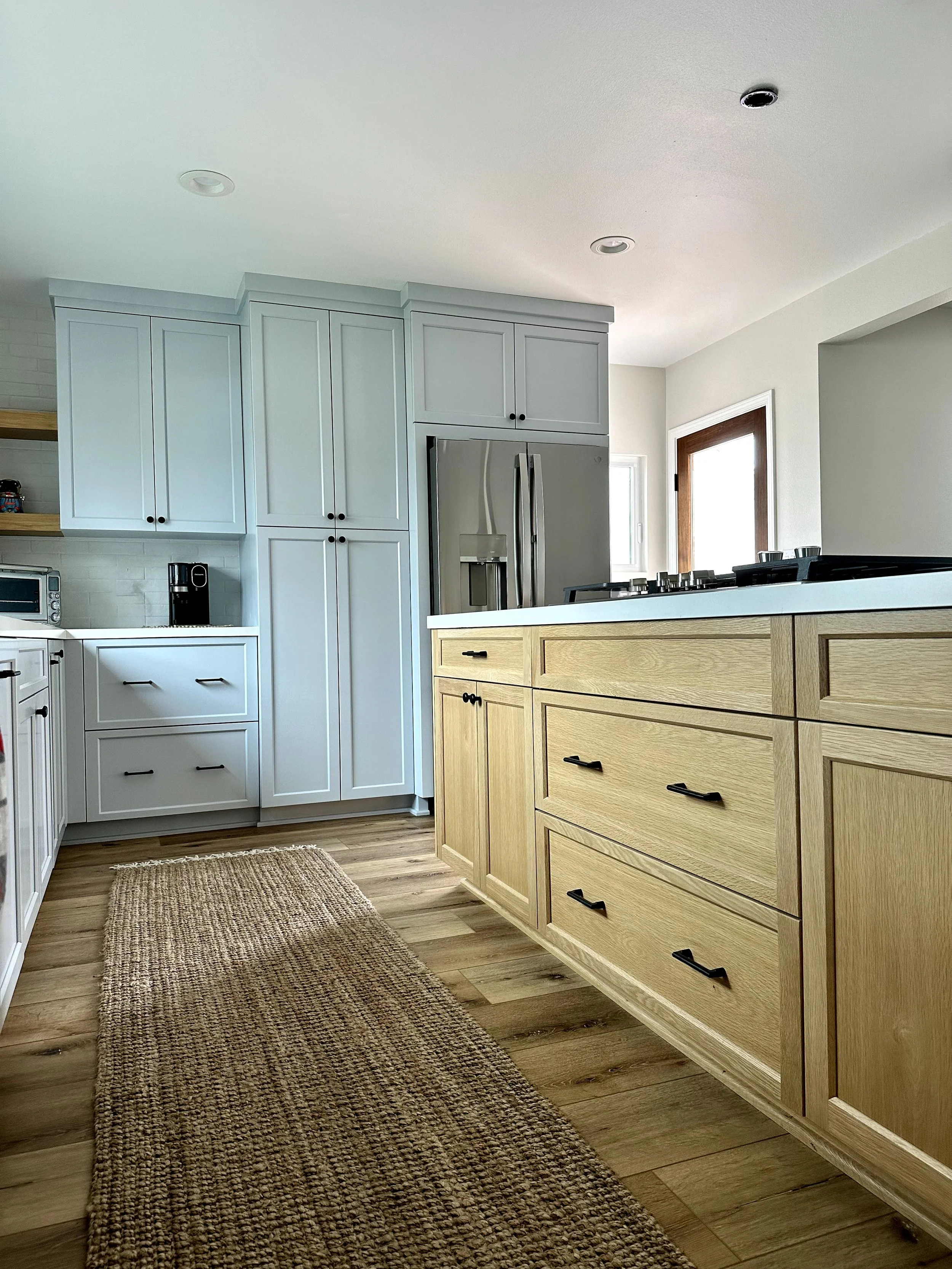 A modern kitchen with white and wood cabinets, stainless steel refrigerator, and a brown woven rug on light wood flooring.