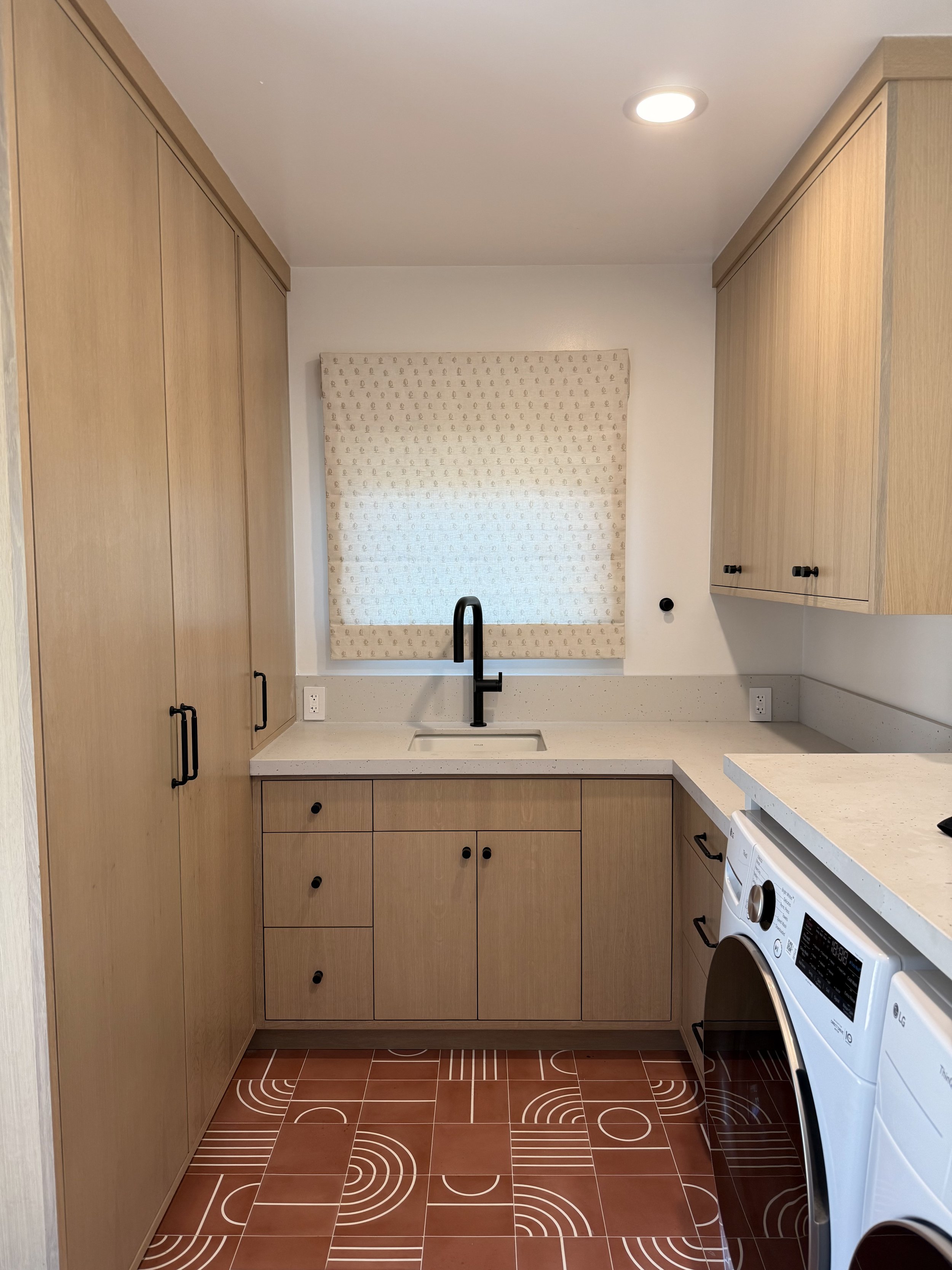 Custom laundry room featuring white oak cabinetry, black hardware, white countertops, and patterned terracotta tile flooring.