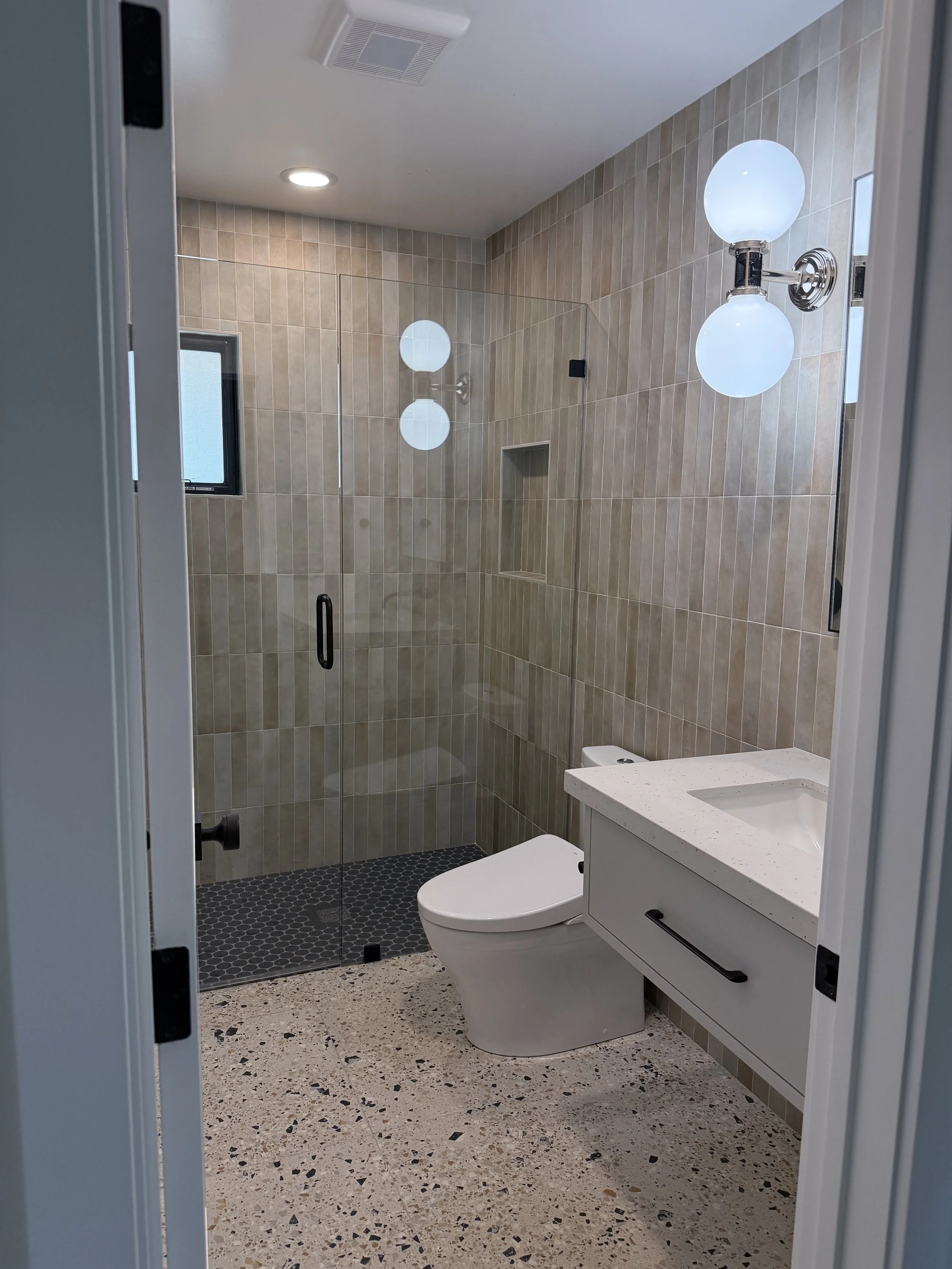Bathroom with dimensional neutral wall tile, terrazzo-style flooring, white vanity, and modern globe lighting.