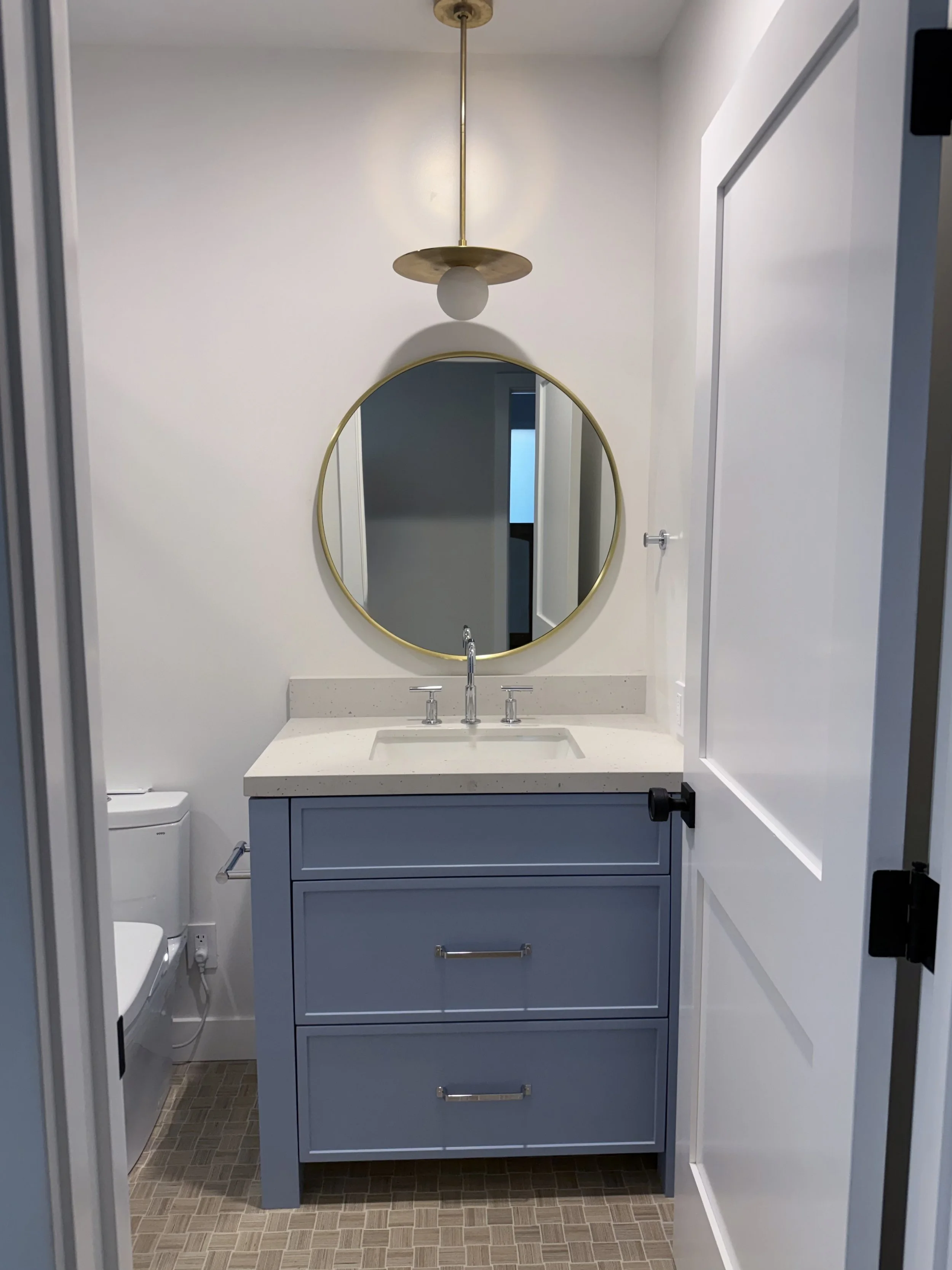 Blue bathroom vanity with white countertop, round brass-framed mirror, and brass wall sconce.