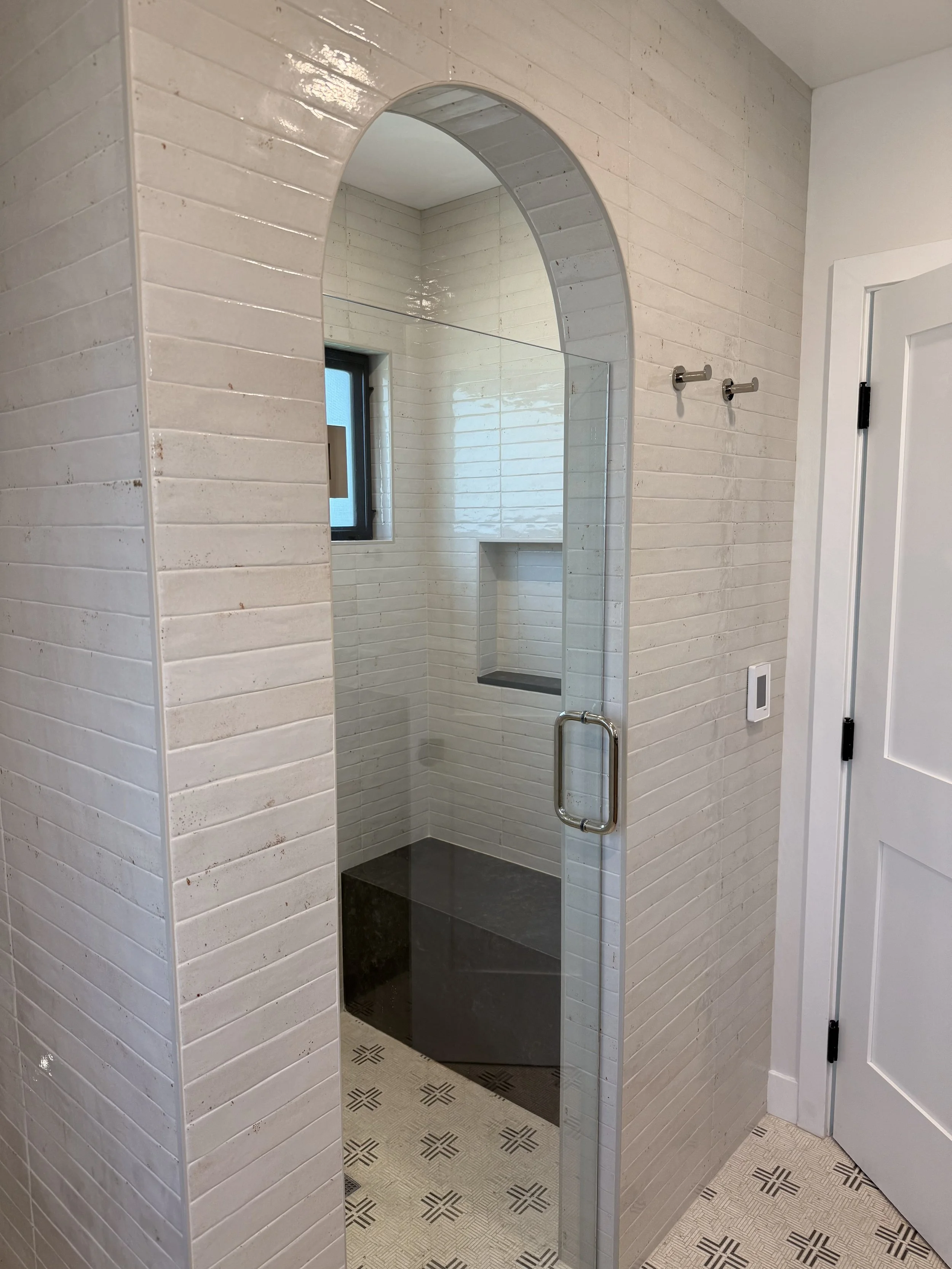 Walk-in shower with arched opening, glass enclosure, neutral tile, built-in shower niche, and dark stone shower floor.