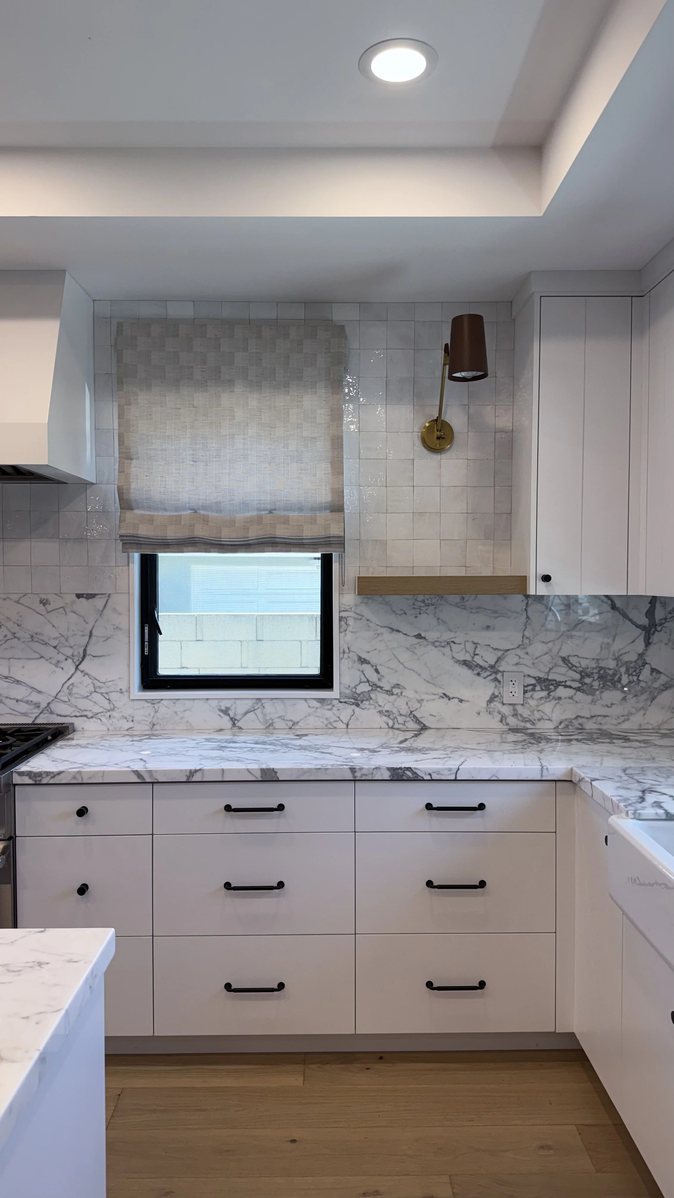 White kitchen cabinetry with natural stone countertop and full-height stone backsplash, accented by a brass wall sconce and Roman shade.