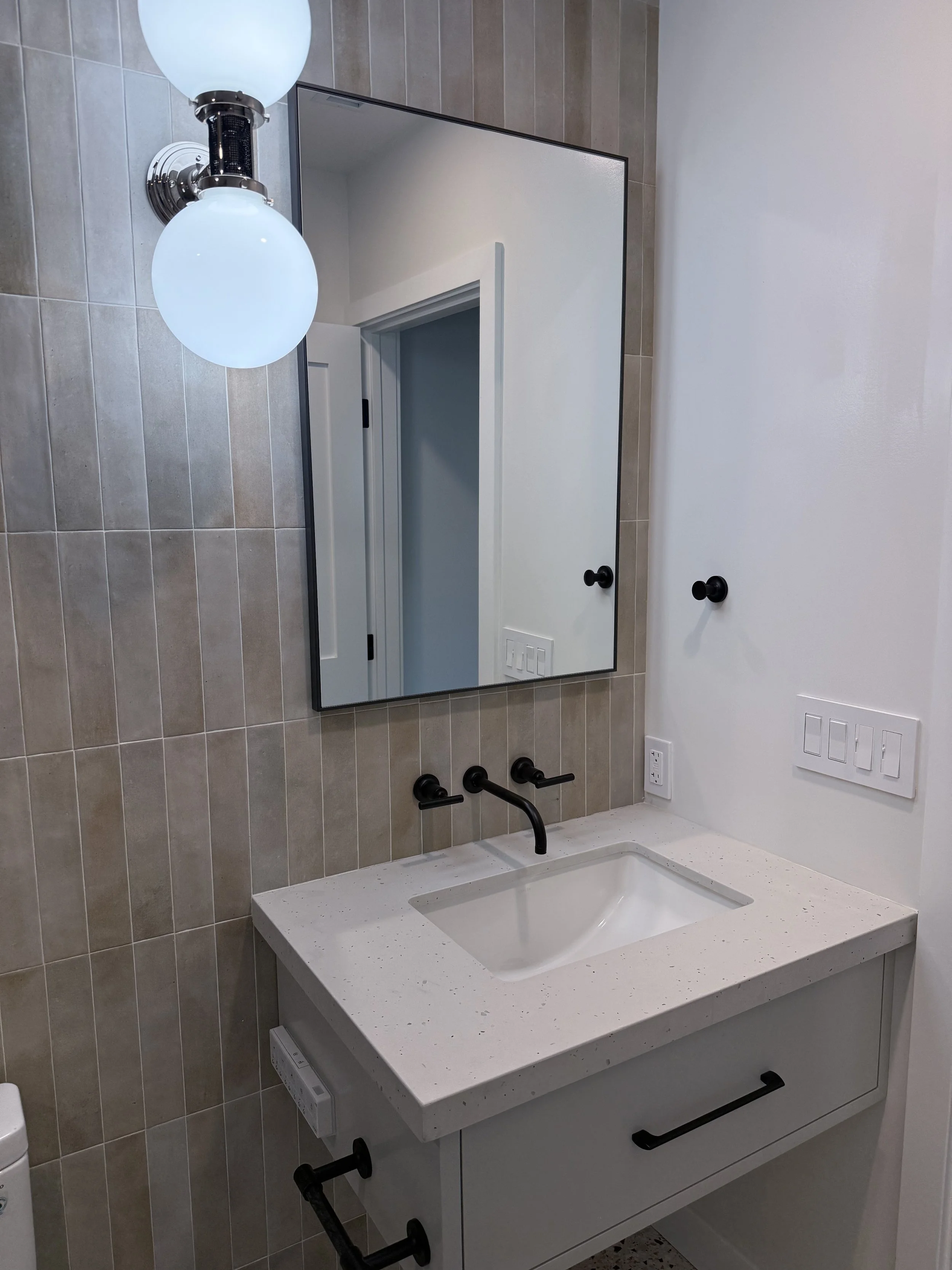 Bathroom vanity with white countertop, undermount sink, black wall-mounted faucet, and handmade-look neutral wall tile.