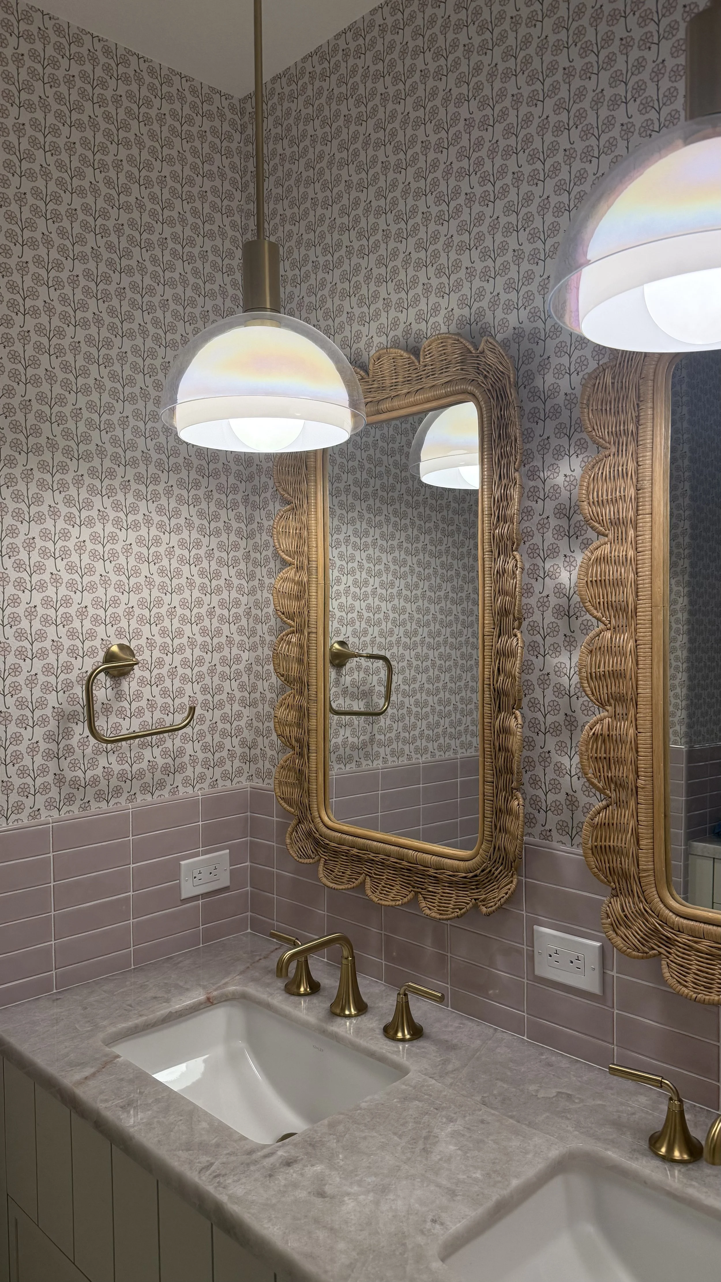 Bathroom with pink tiles and floral wallpaper, featuring two wicker bordered mirrors, gold fixture, and two undermount sinks.