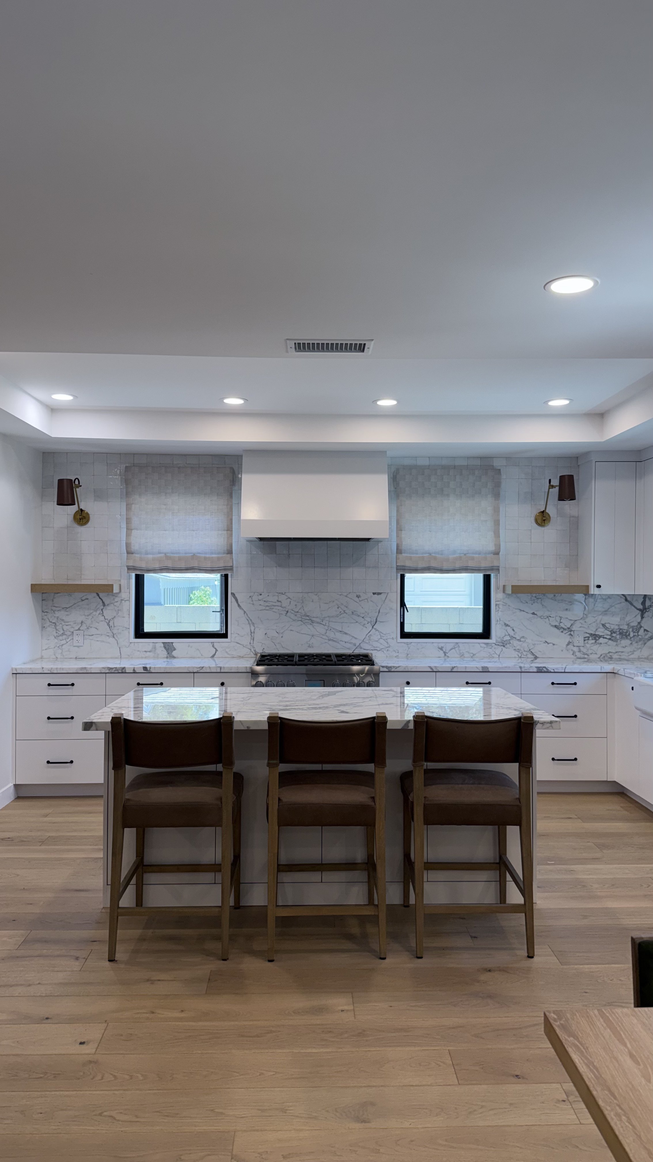 Full view of the remodeled kitchen with white custom cabinetry, stone waterfall island, wood counter stools, and light wood flooring.