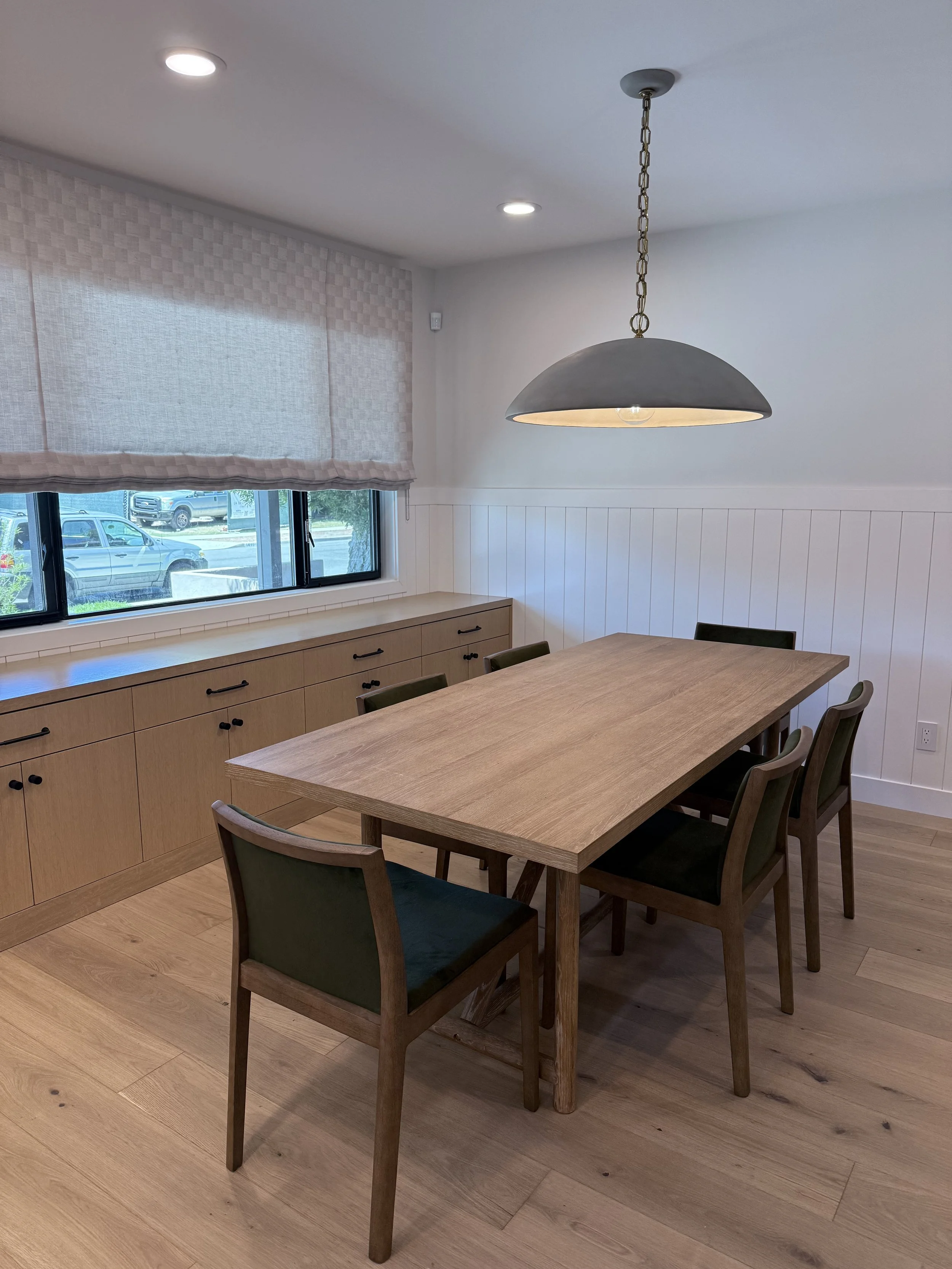 Dining area with custom white oak built-in cabinetry, wood dining table, upholstered chairs, Roman shade, and statement pendant light.