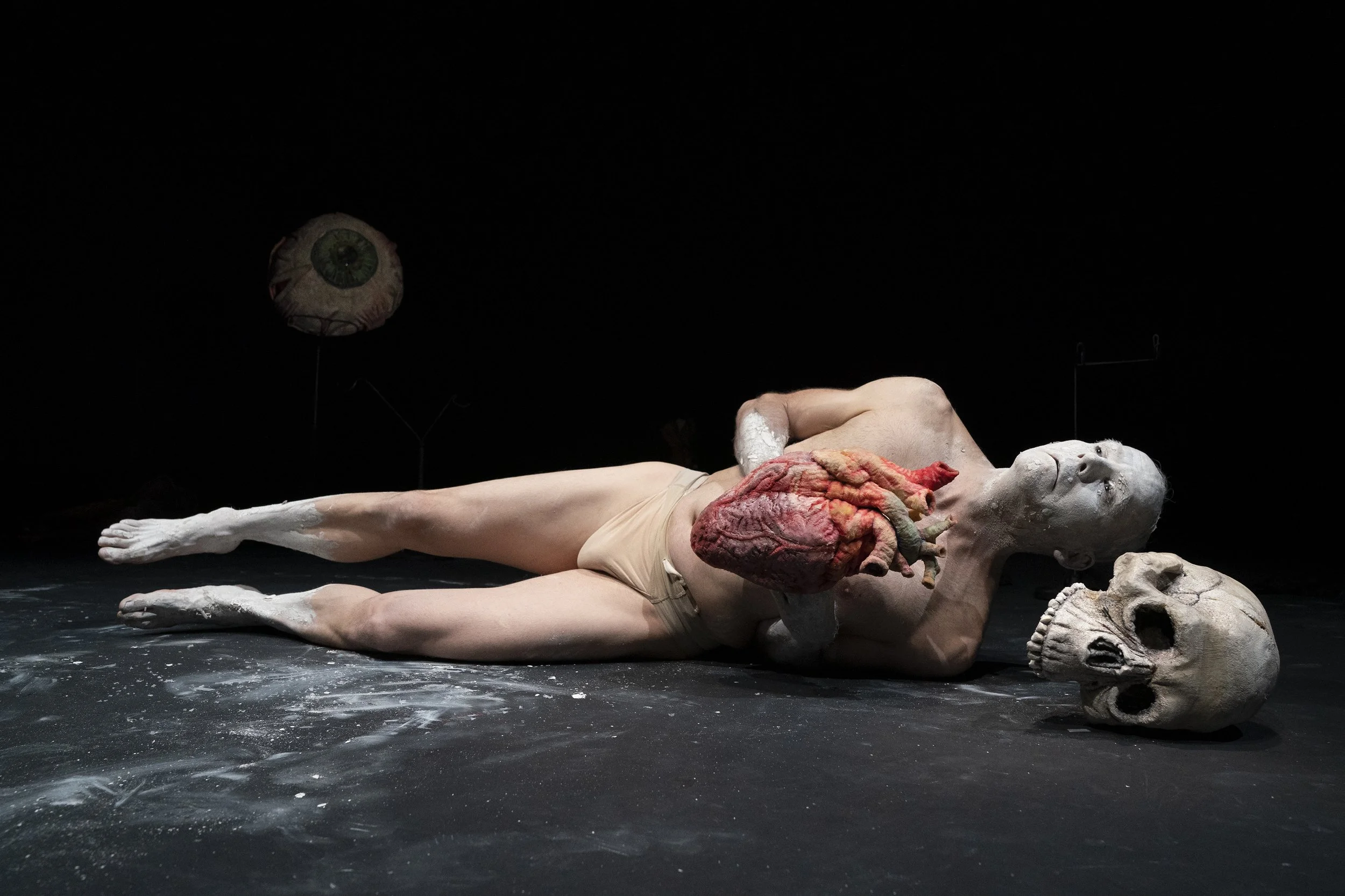 Puppeteer Kevin Augustine from Lone Wolf Tribe lays on stage with face painted white, holding a carved foam heart and surrounded by a foam skull and floating foam eye
