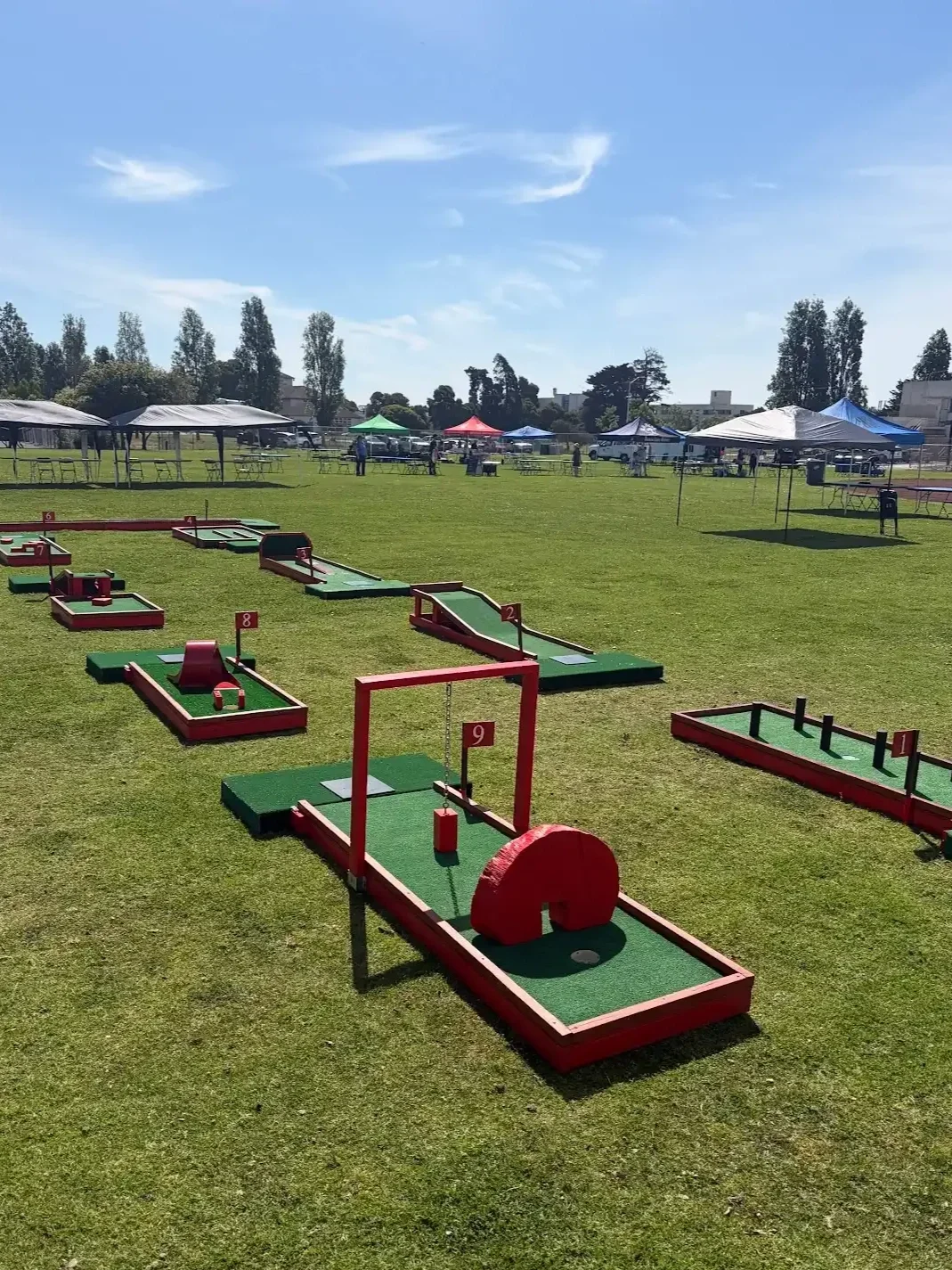 Miniature golf course set up for fund raiser entertainment and activity on a grassy field with tents and people in the background under a blue sky with a few clouds.