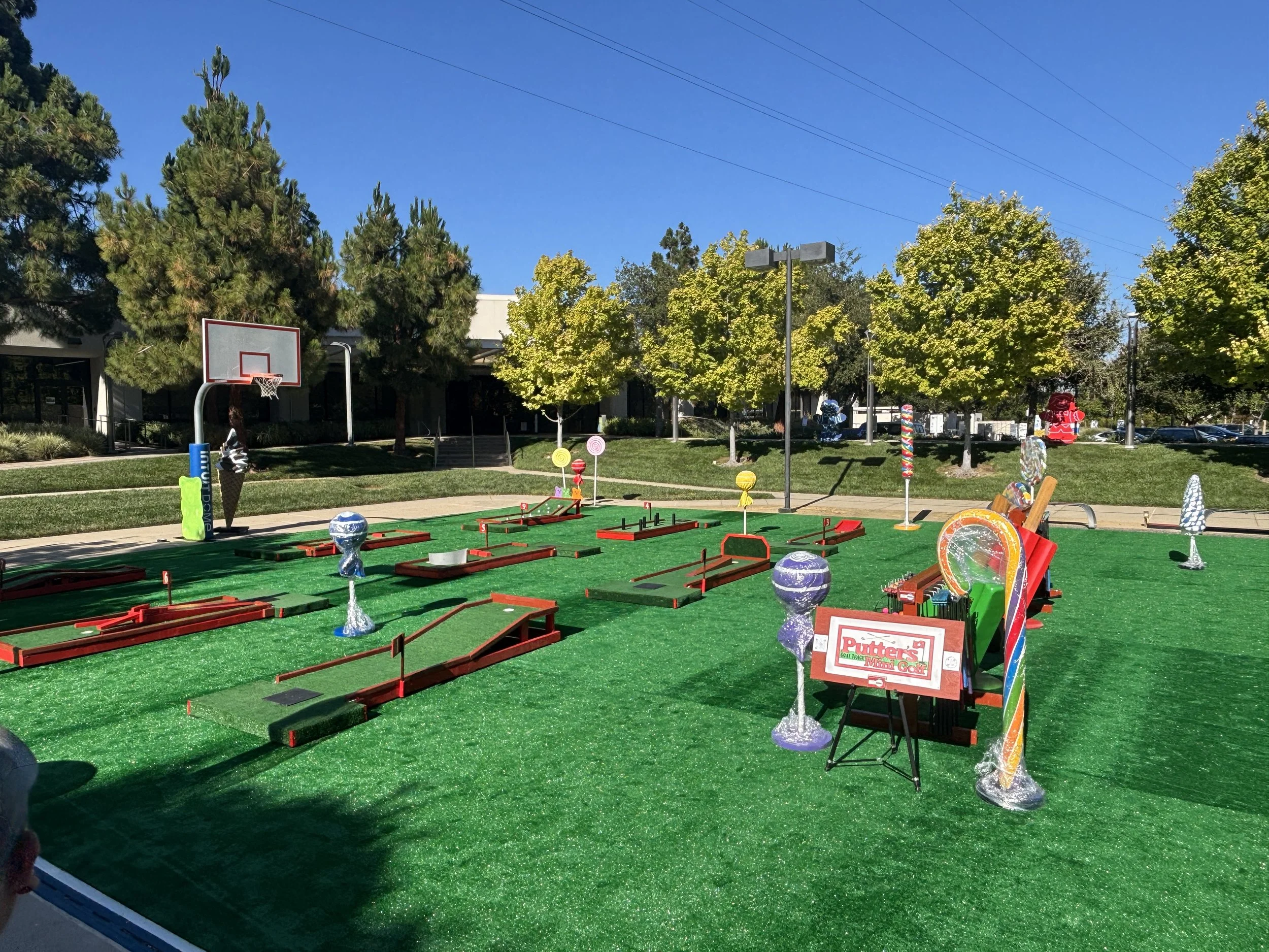 Miniature golf course with various obstacles, colorful decorations, and a sign that reads 'Putter's Mini Golf' in a park setting with trees and a clear blue sky. Rental miniature golf set up for functions and events.