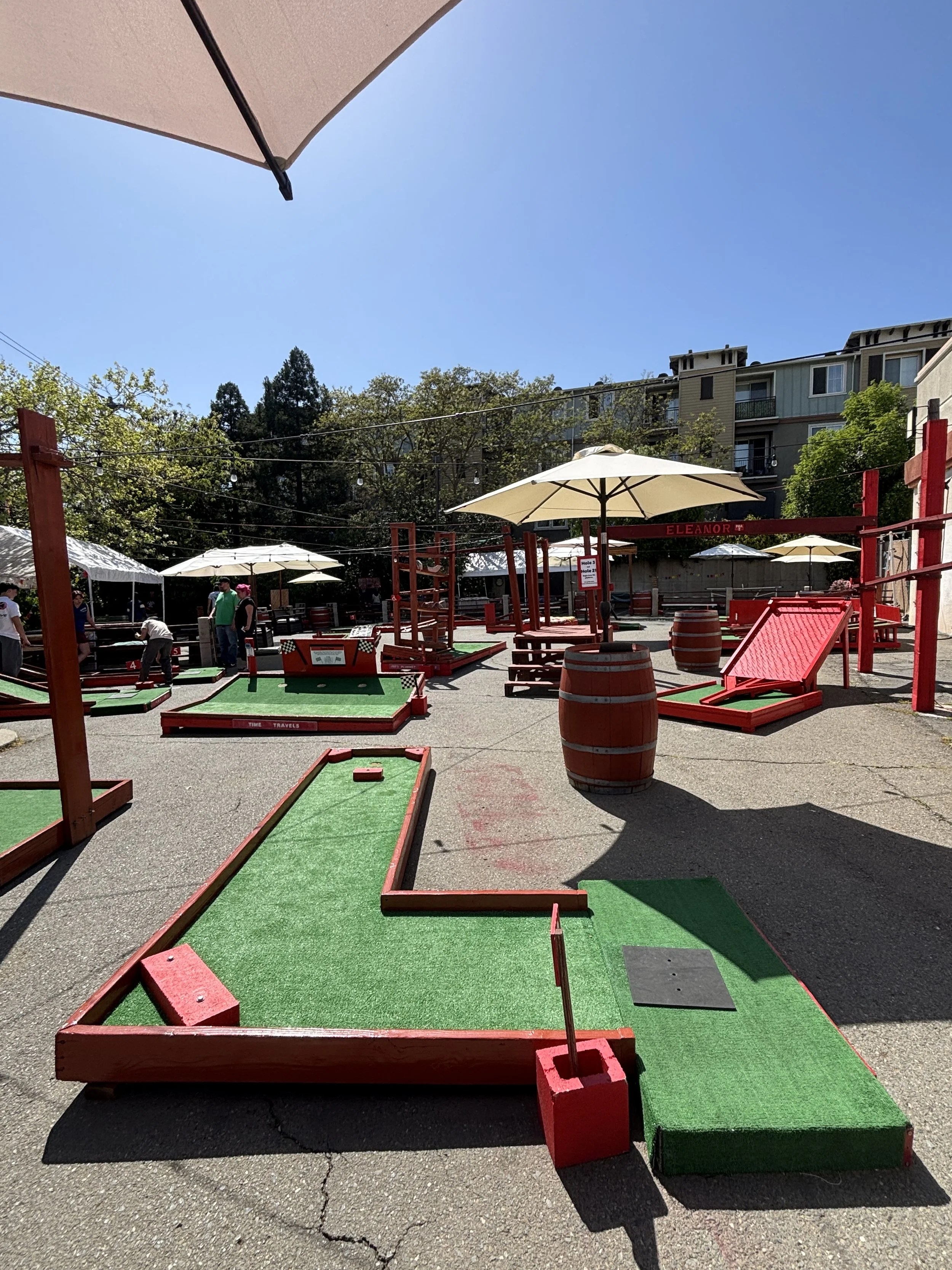 Outdoor mini golf course with red wooden obstacles, green artificial turf, barrel decor, and patio umbrellas under a clear sky.