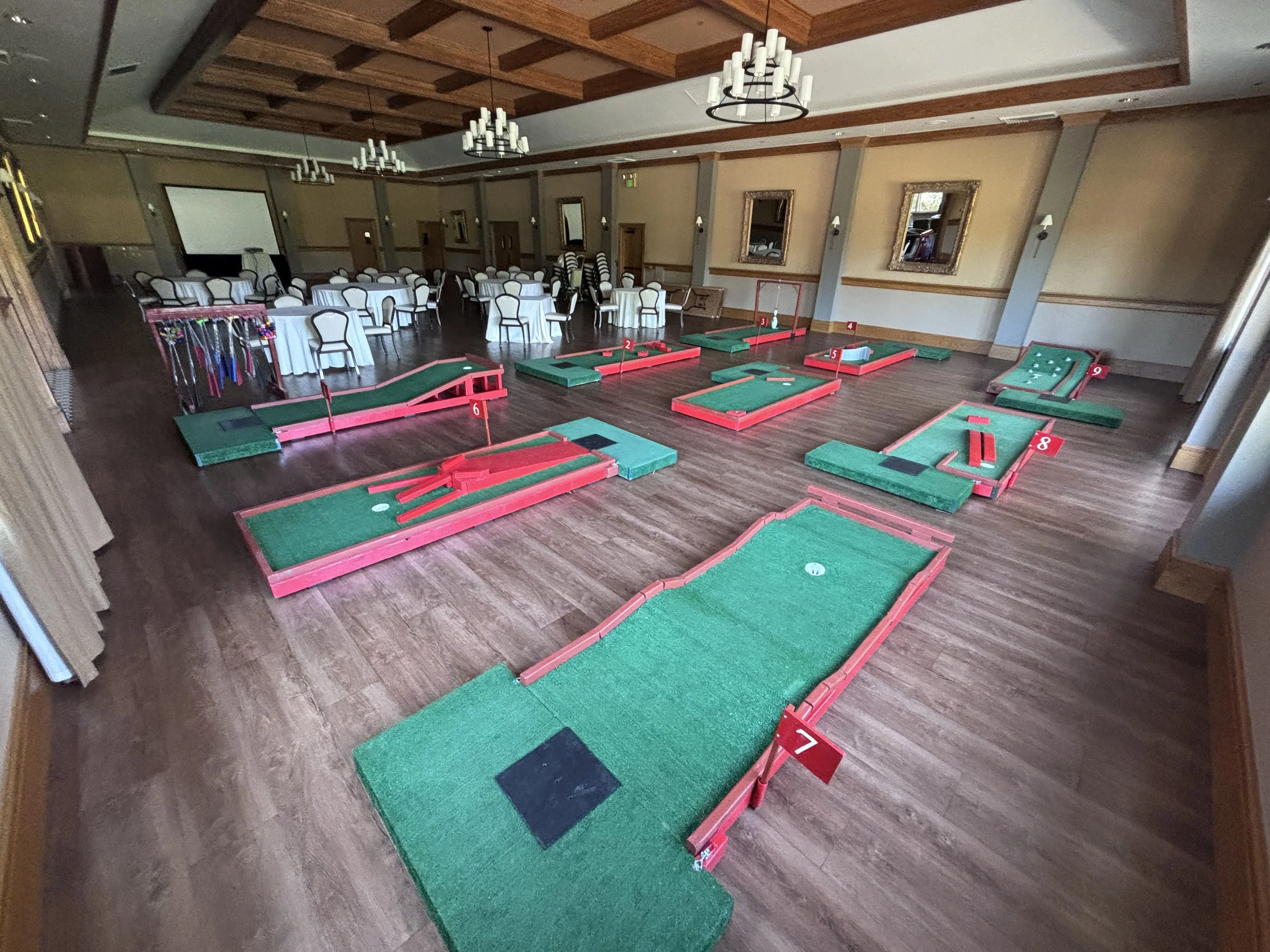 Indoor mini golf course available for delivery with 3 to 18 holes with unique obsticles and barriers, set up in a banquet hall with round tables and chairs.