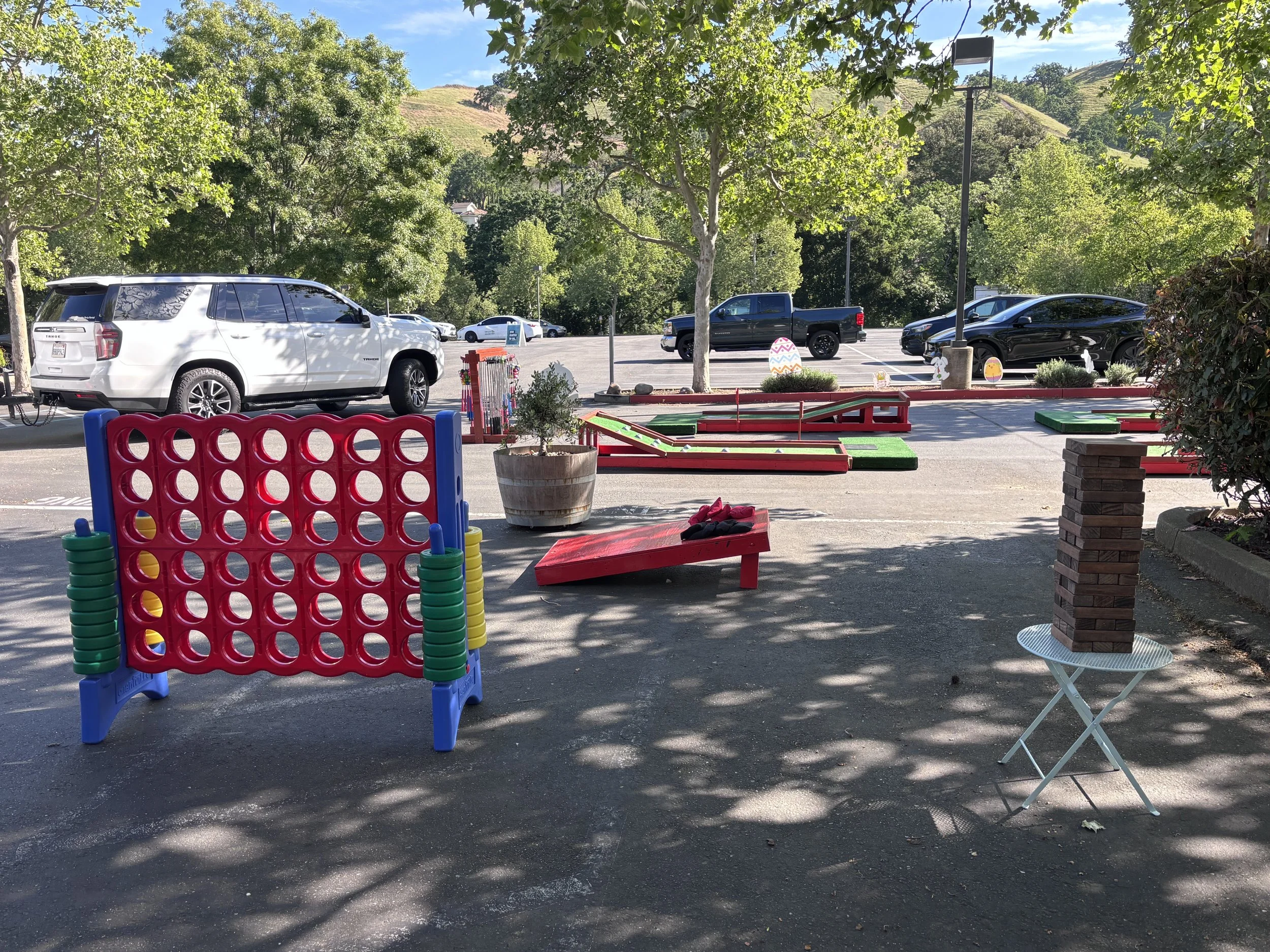 A parking lot seen from a shaded outdoor area with trees and hills in the background. Several parked cars, including a white SUV and a black pickup truck, are visible. In the foreground, there is a red and blue Connect Four game, a large plant pot, a