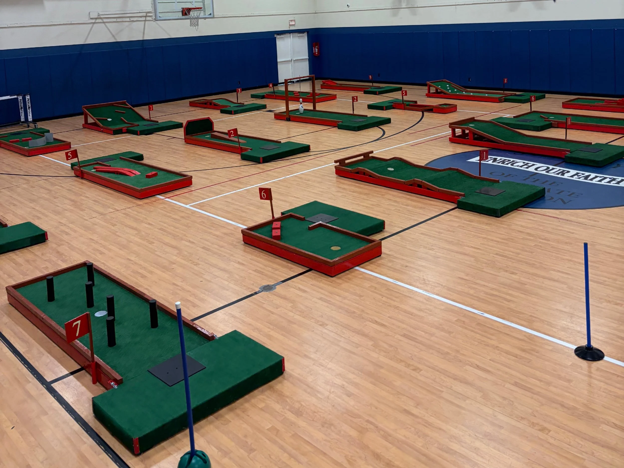 Indoor mini golf course with multiple holes, flags, and obstacles on a basketball court with wooden flooring.