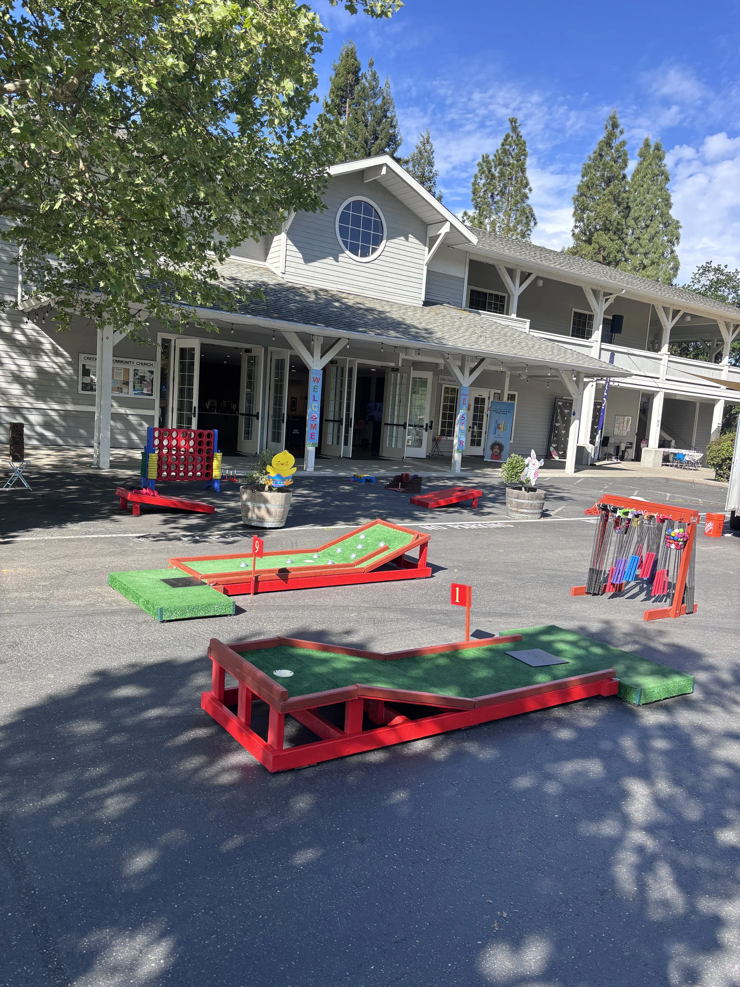 Outdoor scene with mini golf course setup in front of a multi-story building, surrounded by trees and clear blue sky.