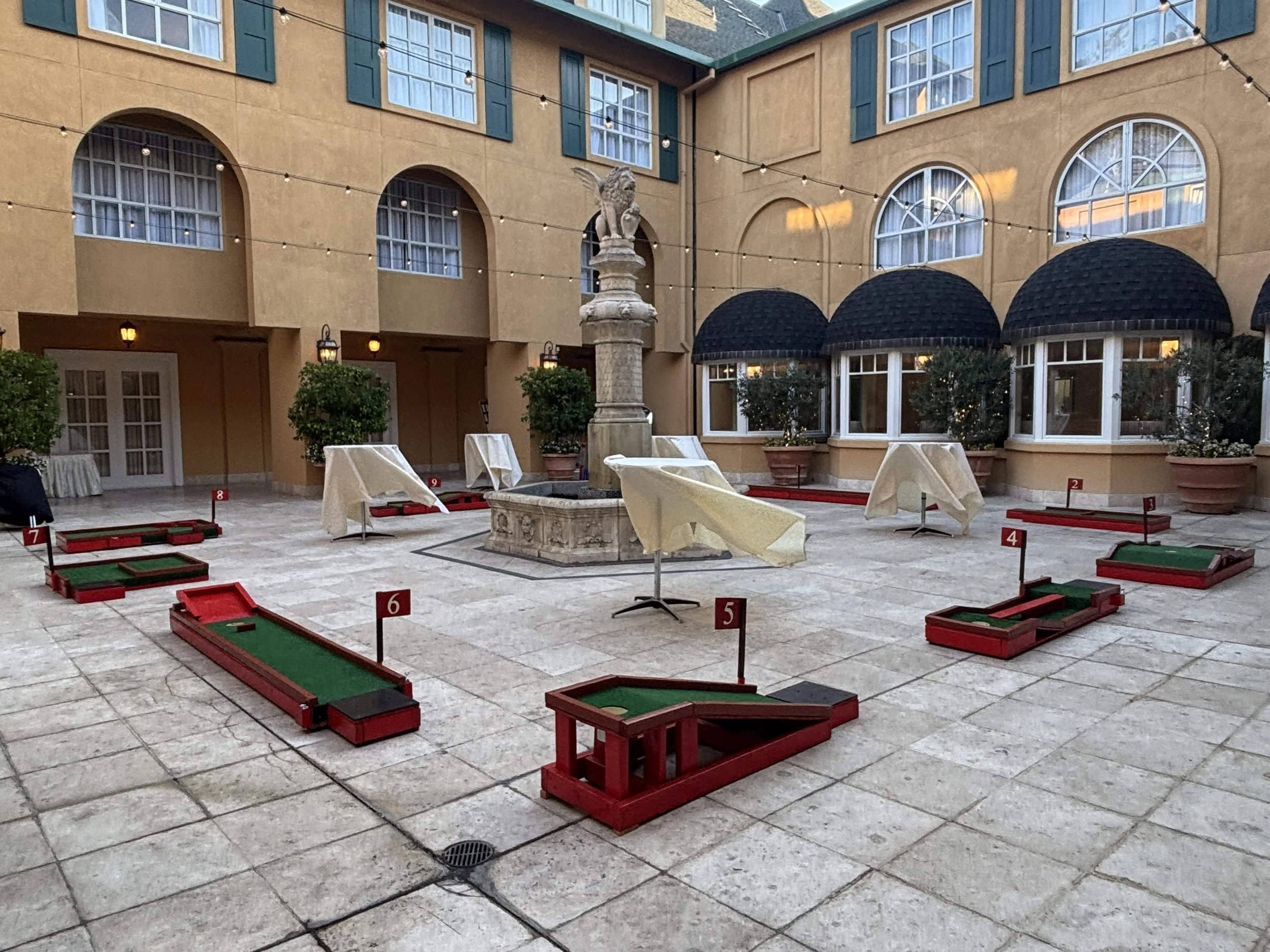 Miniature golf delivered for a courtyard wedding with string lights, a stone statue fountain of a mythical creature, and mini golf course obstacles with numbered markers.