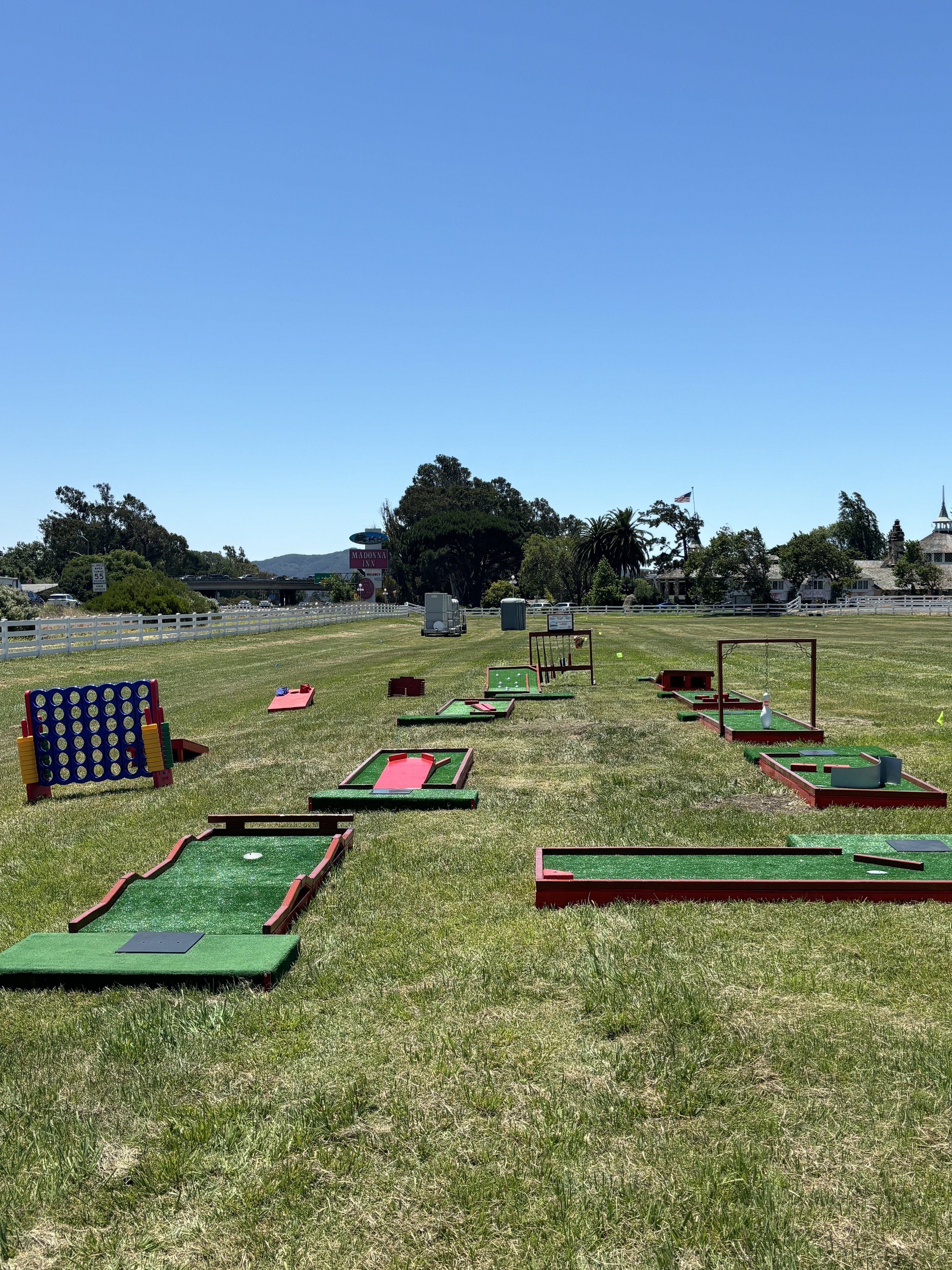 Mini-golf course on grass with multiple holes and obstacles, outdoor setting, clear blue sky, trees in background.