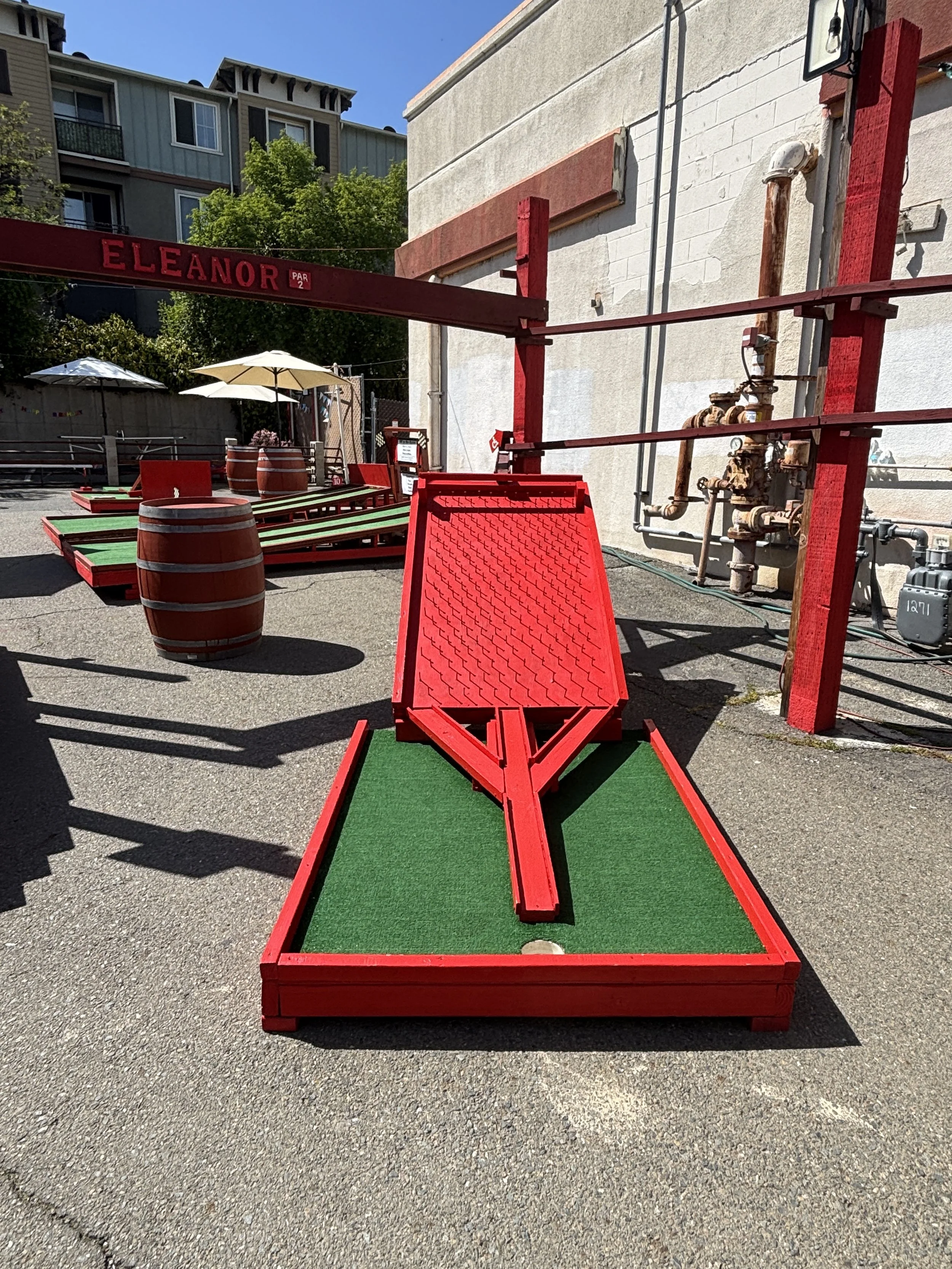 Outdoor mini-golf course with a red obstacle named Eleanor, featuring red wooden structures, green artificial turf, and barrel decorations.