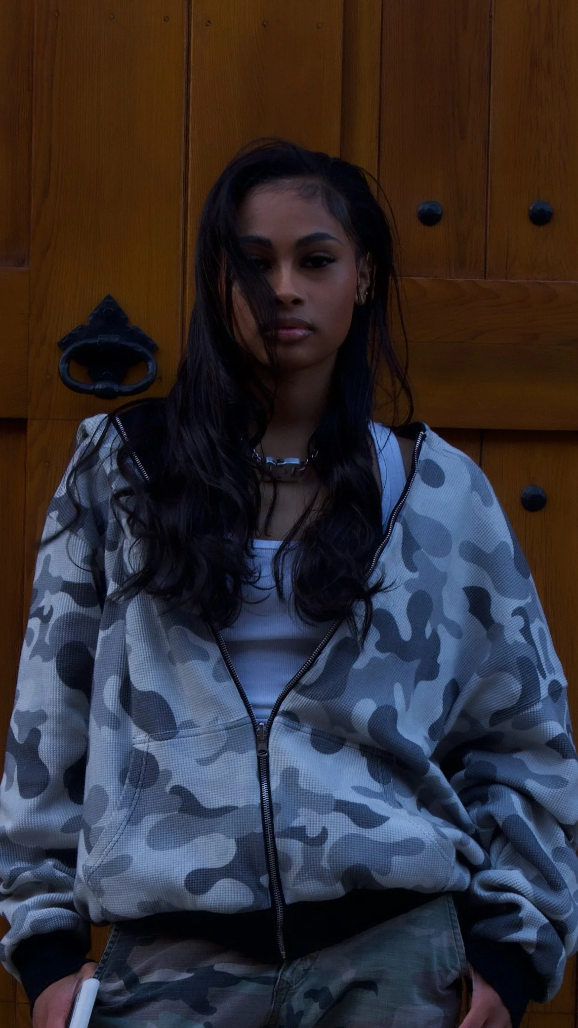 Young woman with long dark hair wearing a camouflage-patterned jacket, white top, and chunky chain necklace, standing in front of wooden cabinets.