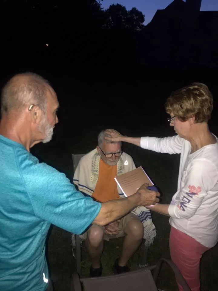 Three people outdoors at dusk; one man sitting with a book and prayer shawl, a man and woman touching his head blessing him