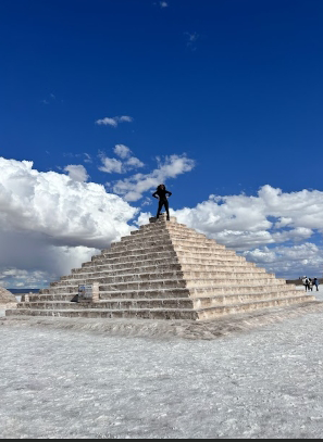 A person standing on top of a large stone pyramid with a bright blue sky and clouds in the background.